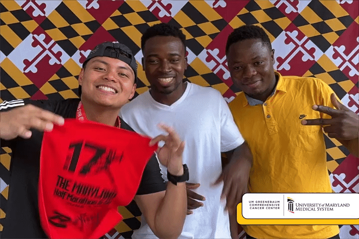 Three smiling men posing together indoors in front of a Maryland-themed background. The man on the left is holding a red piece of cloth with black text, wearing a black cap and shirt, making a peace sign. The man in the middle is wearing a white shirt, smiling. The man on the right is wearing a yellow shirt, also making a peace sign.