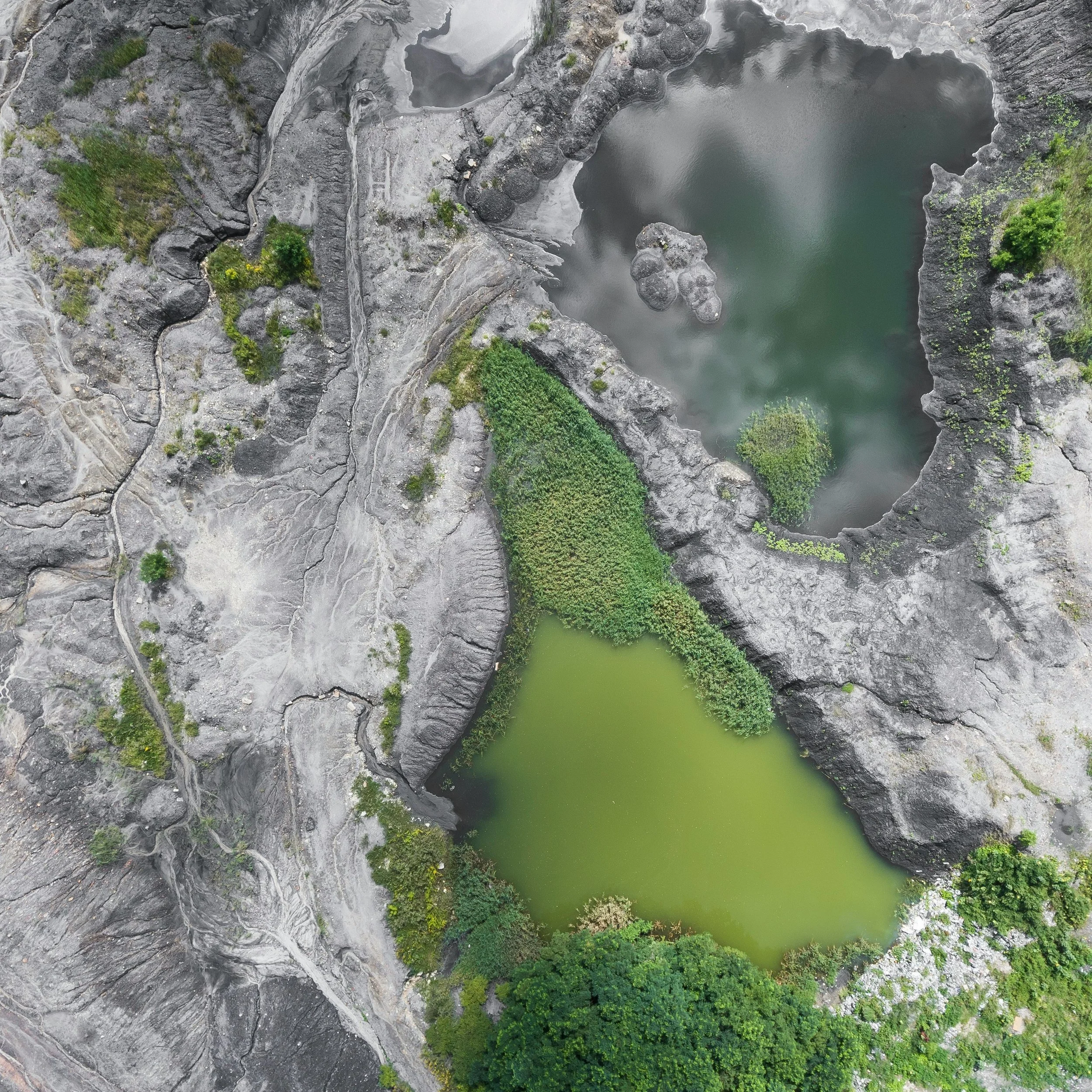 Aerial view of a volcanic crater with two lakes, one dark and one green, surrounded by rocky terrain and sparse greenery.