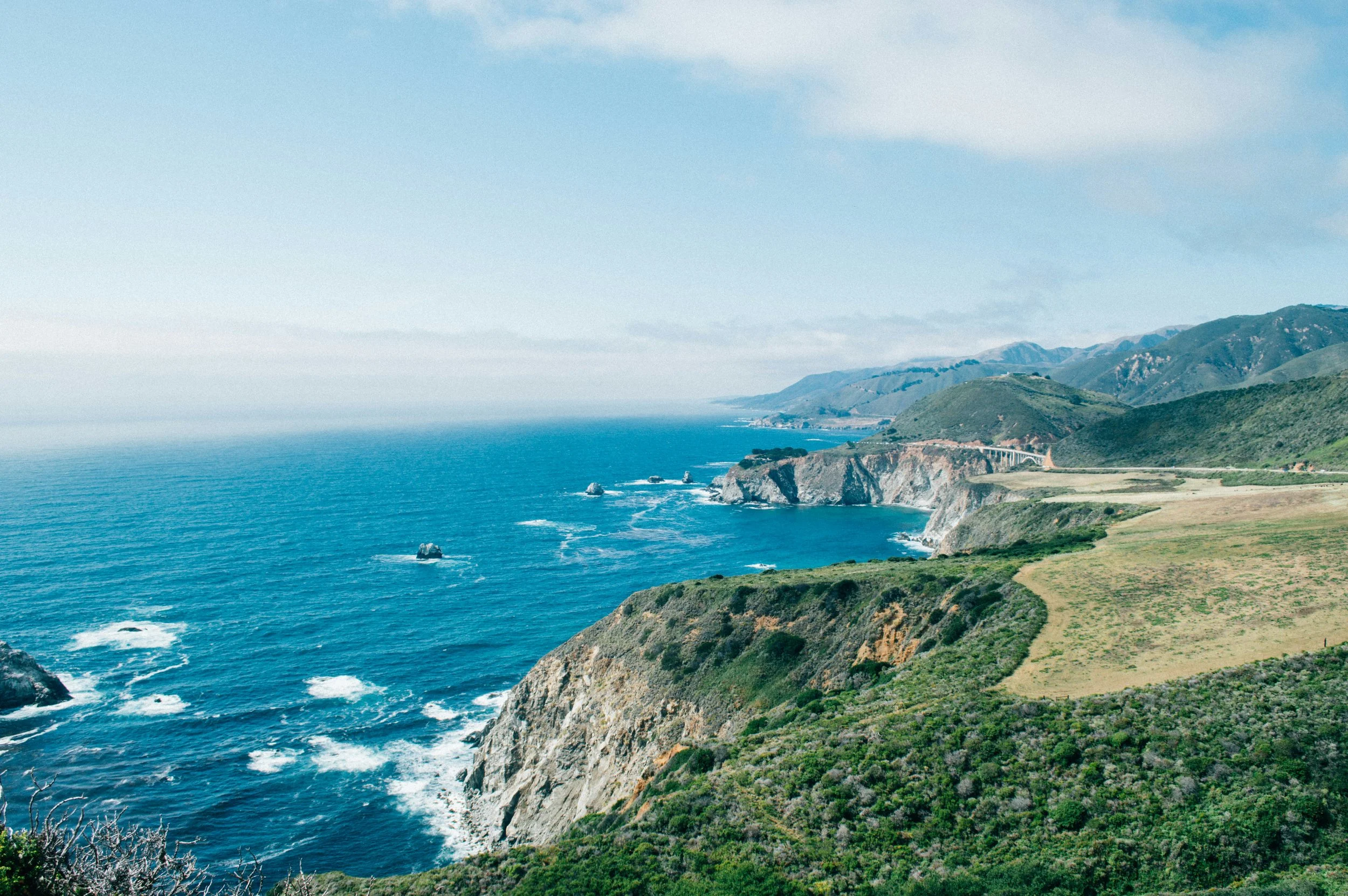 Scenic view of steep coastal cliffs with green vegetation, blue ocean with small rocks and waves, and hills in the background under a partly cloudy sky.