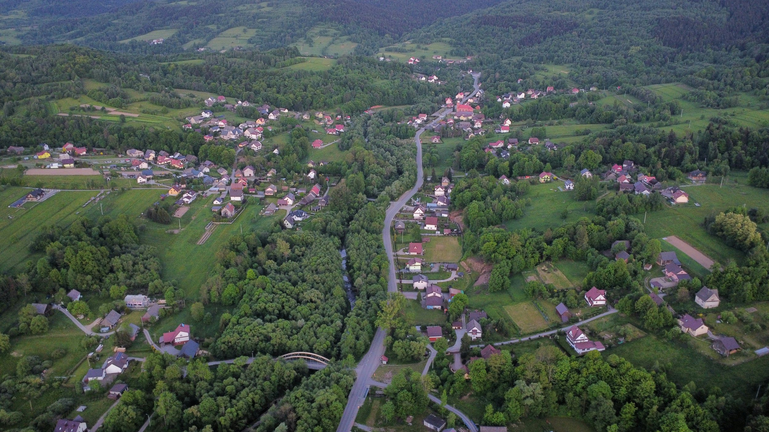 Aerial view of a small town with houses, roads, and dense greenery, surrounded by hills.
