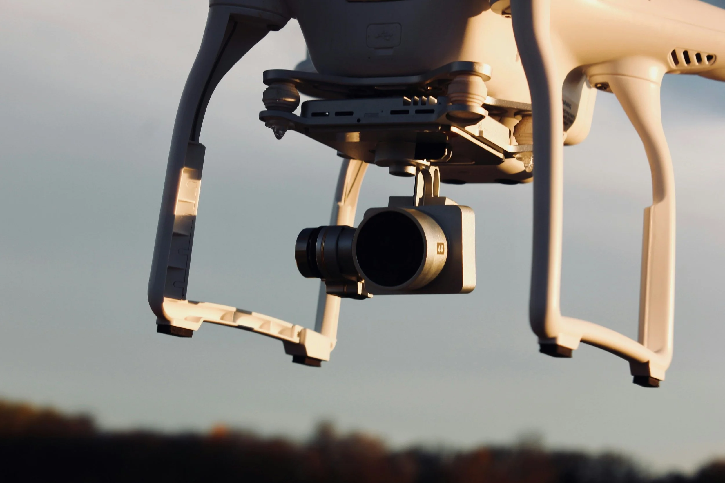A close-up view of a white quadcopter drone with a mounted 4K camera, flying outdoors during sunset or sunrise.