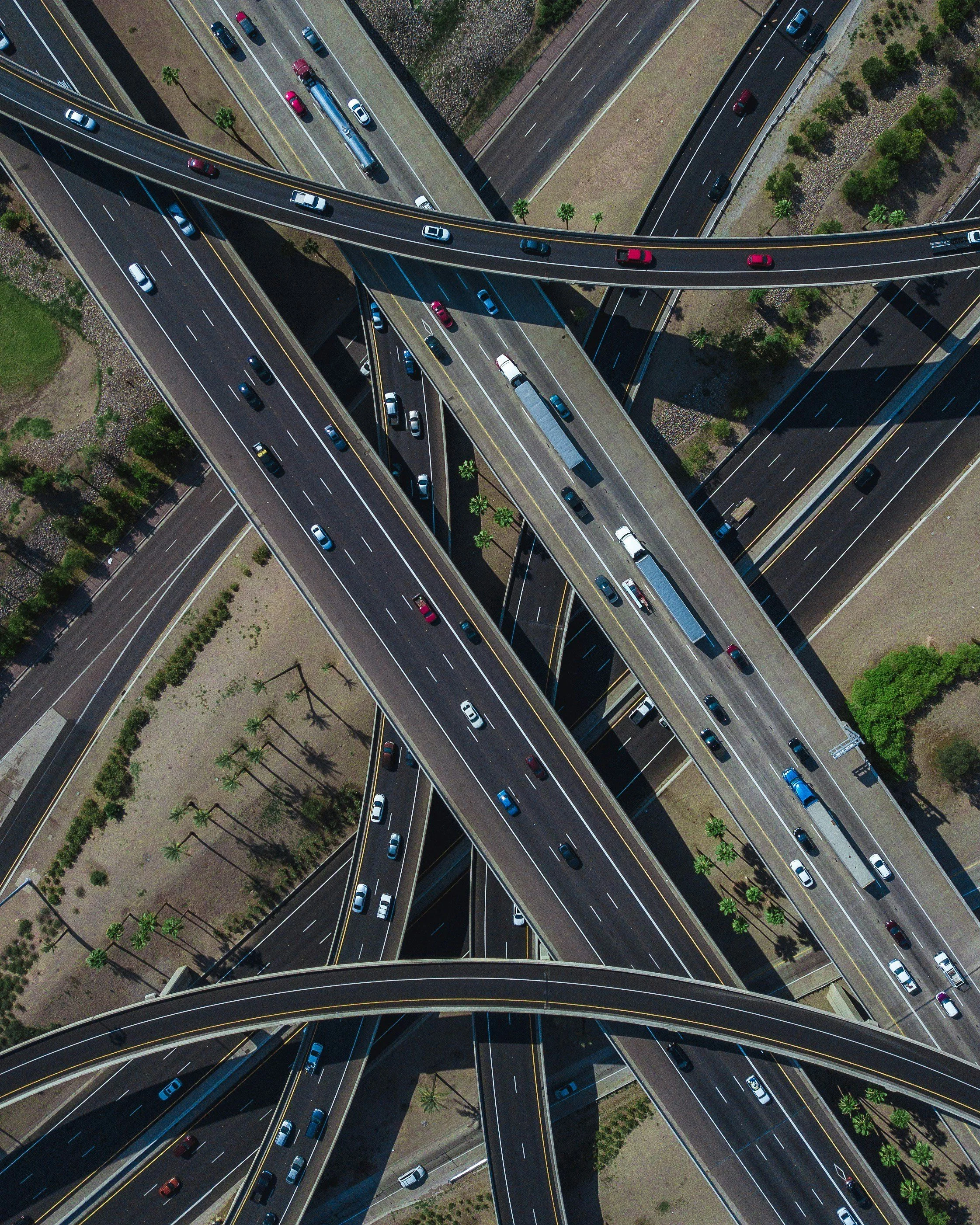 An aerial view of multiple highways and overpasses with cars traveling in various directions. The roads intersect and cross above and below each other, creating a complex highway interchange in a sunny area with patches of green and landscaped areas.