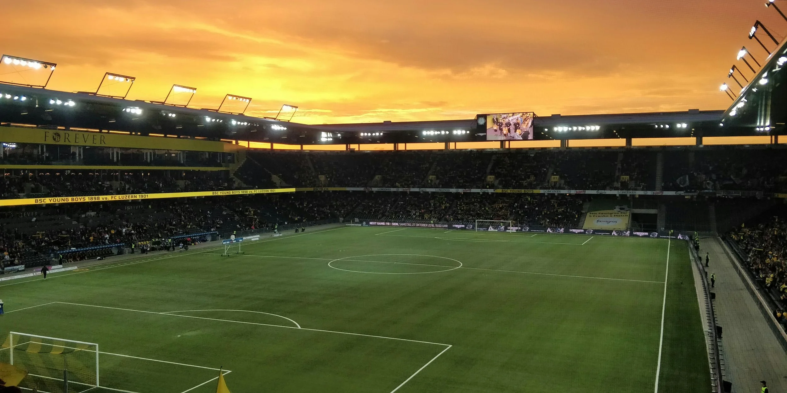 Empty soccer stadium at sunset with vibrant orange sky, illuminated floodlights, and spectators in the stands.