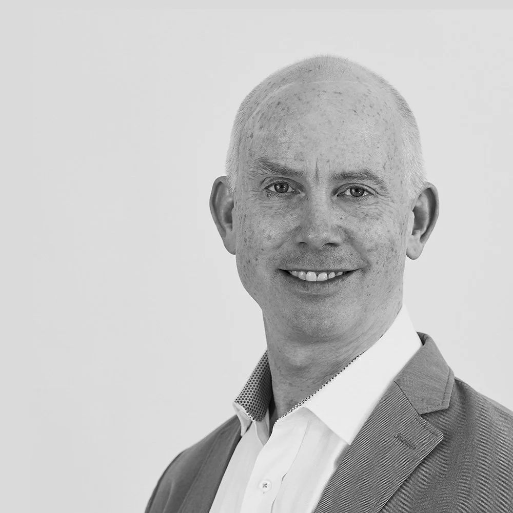 Black and white portrait of Jonathan Posnett, wearing a suit and white shirt, smiling at the camera.