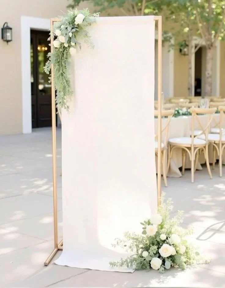 Wedding or event backdrop with white drapery, decorated with white roses and greenery in the top left corner and at the bottom right corner, set outdoors near a building with trees and a table with chairs in the background.