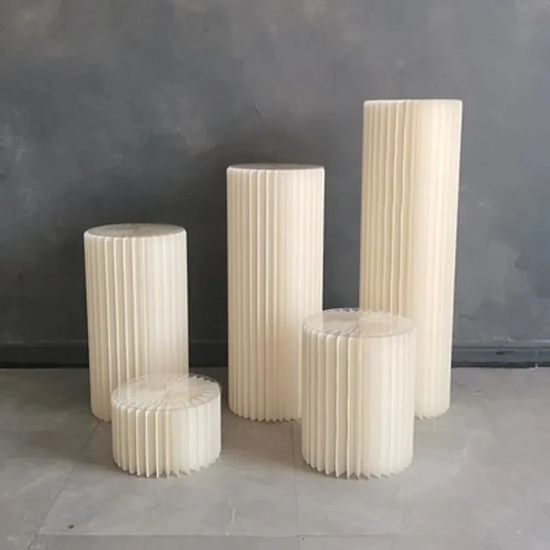 Five cream-colored, vertically pleated cylindrical objects of various heights arranged against a gray wall.