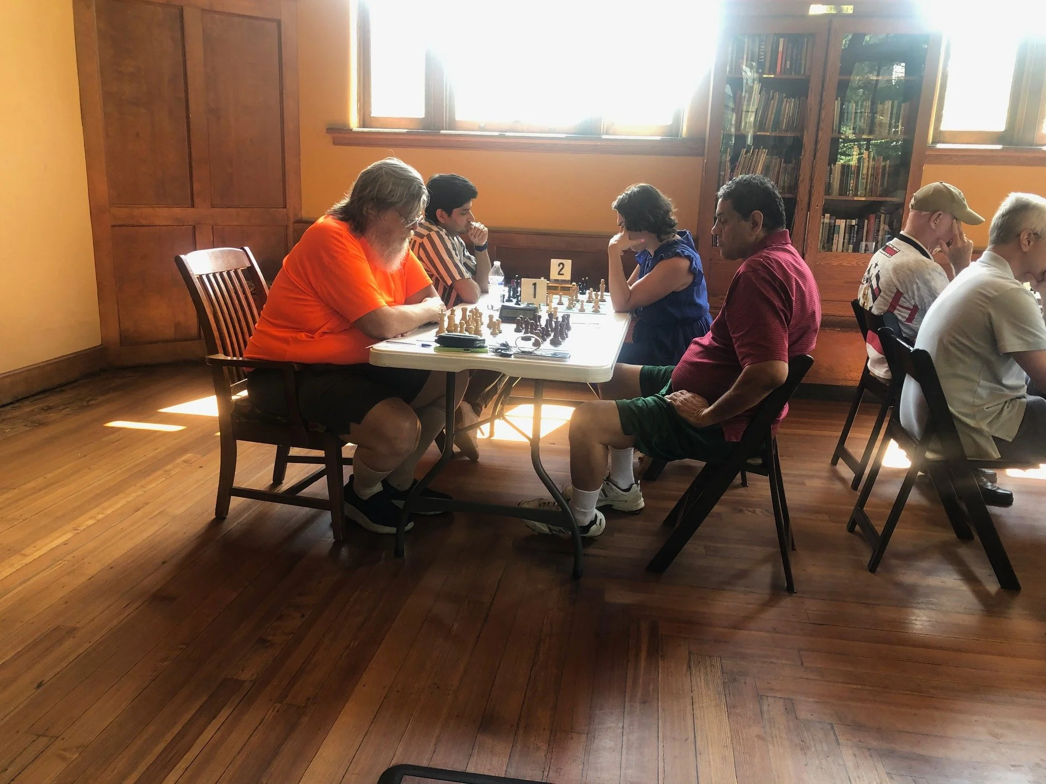 People playing chess at a table in a room with wooden floors, walls, and bookshelves, with sunlight coming through the windows.