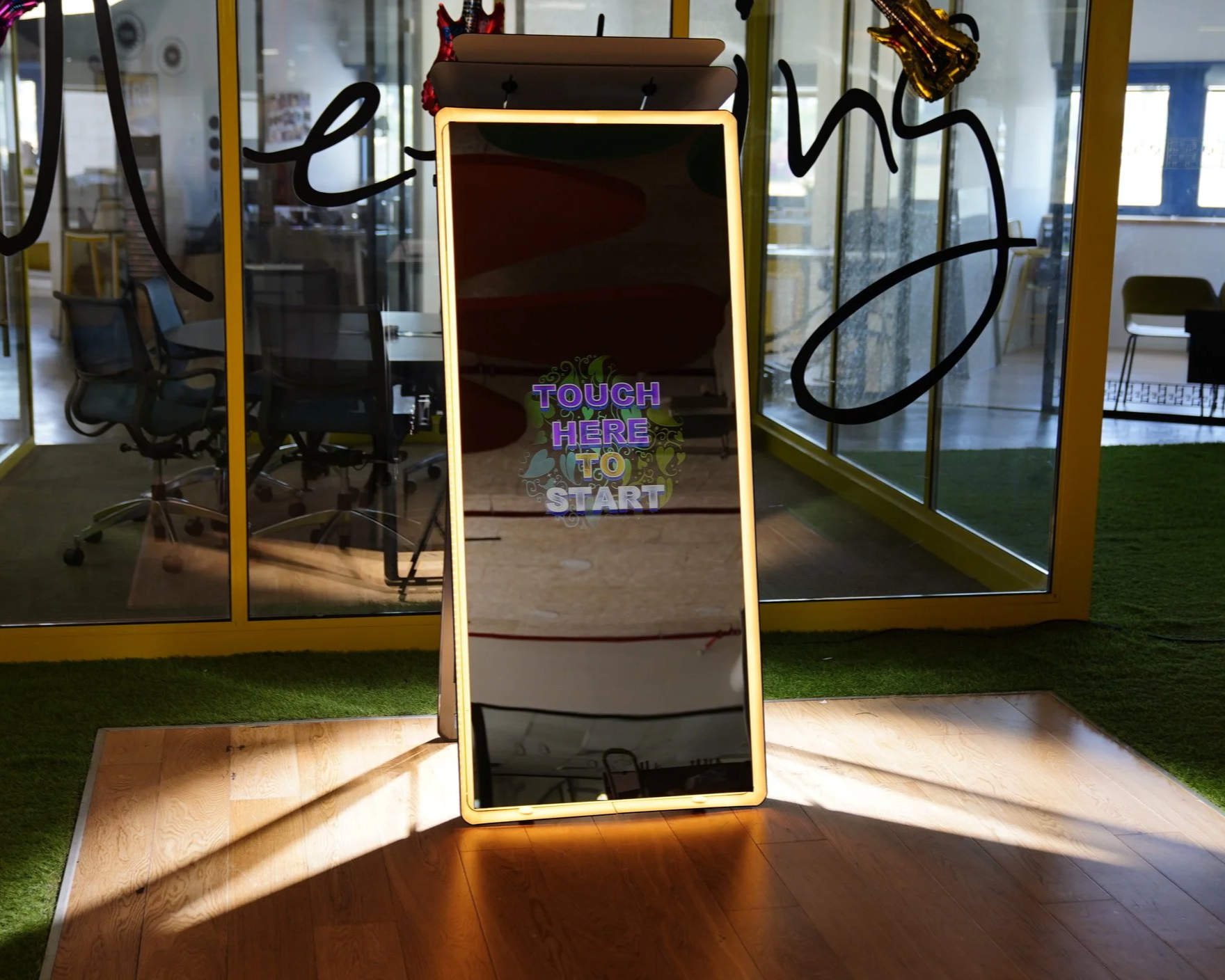 A large vertical digital mirror with a glowing yellow border, displaying the message "Touch Here To Start" in colorful letters. The mirror is placed on a wooden floor, with indoor office chairs and glass walls in the background. Decorative balloons are attached to the top of the mirror.