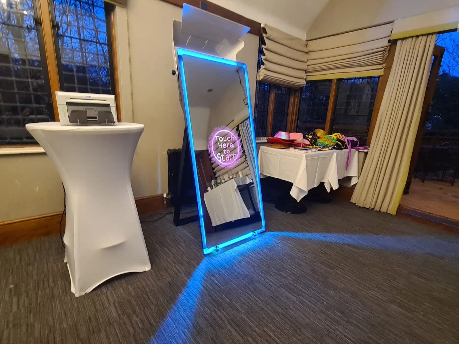 Decorative mirror with blue neon lights and a pink neon sign that says 'Touch Here to Start' leaning against the wall in a room with beige curtains, a table covered with pink hats, colorful hair accessories, and other items, and a high-top table with a small electronic device.