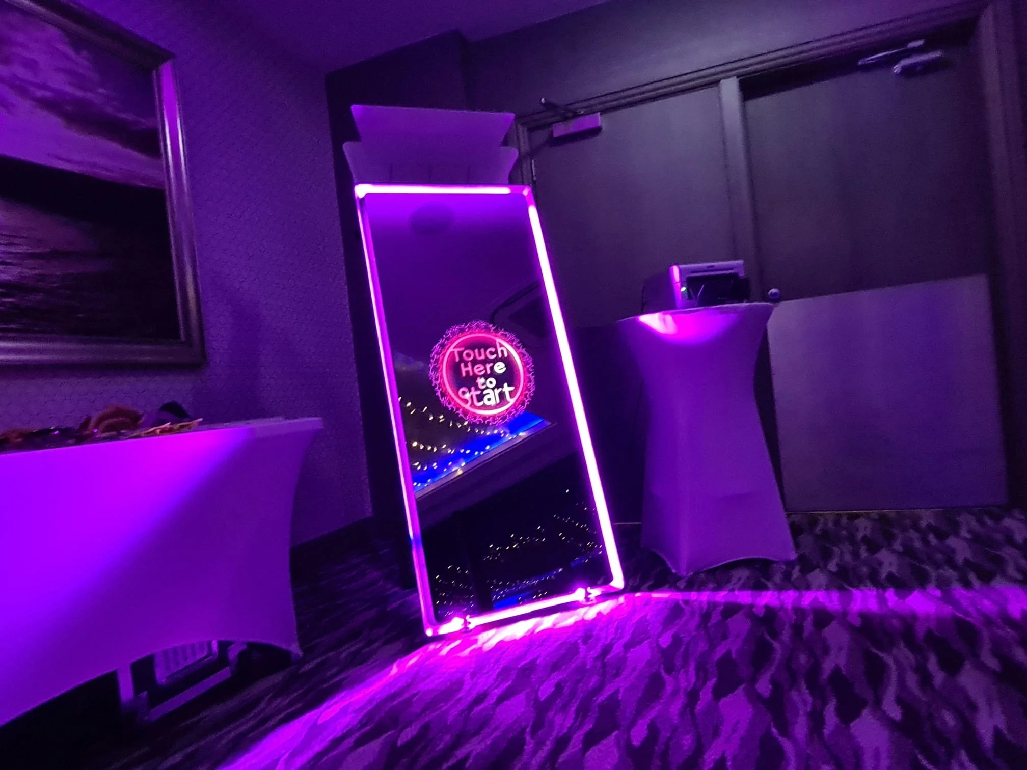 A neon sign that says 'Touch Here to Start' is leaning against the wall in a dimly lit room with purple lighting. In front of the sign, there are two white cocktail tables with some items on top, and the room's floor has a leopard-print carpet.