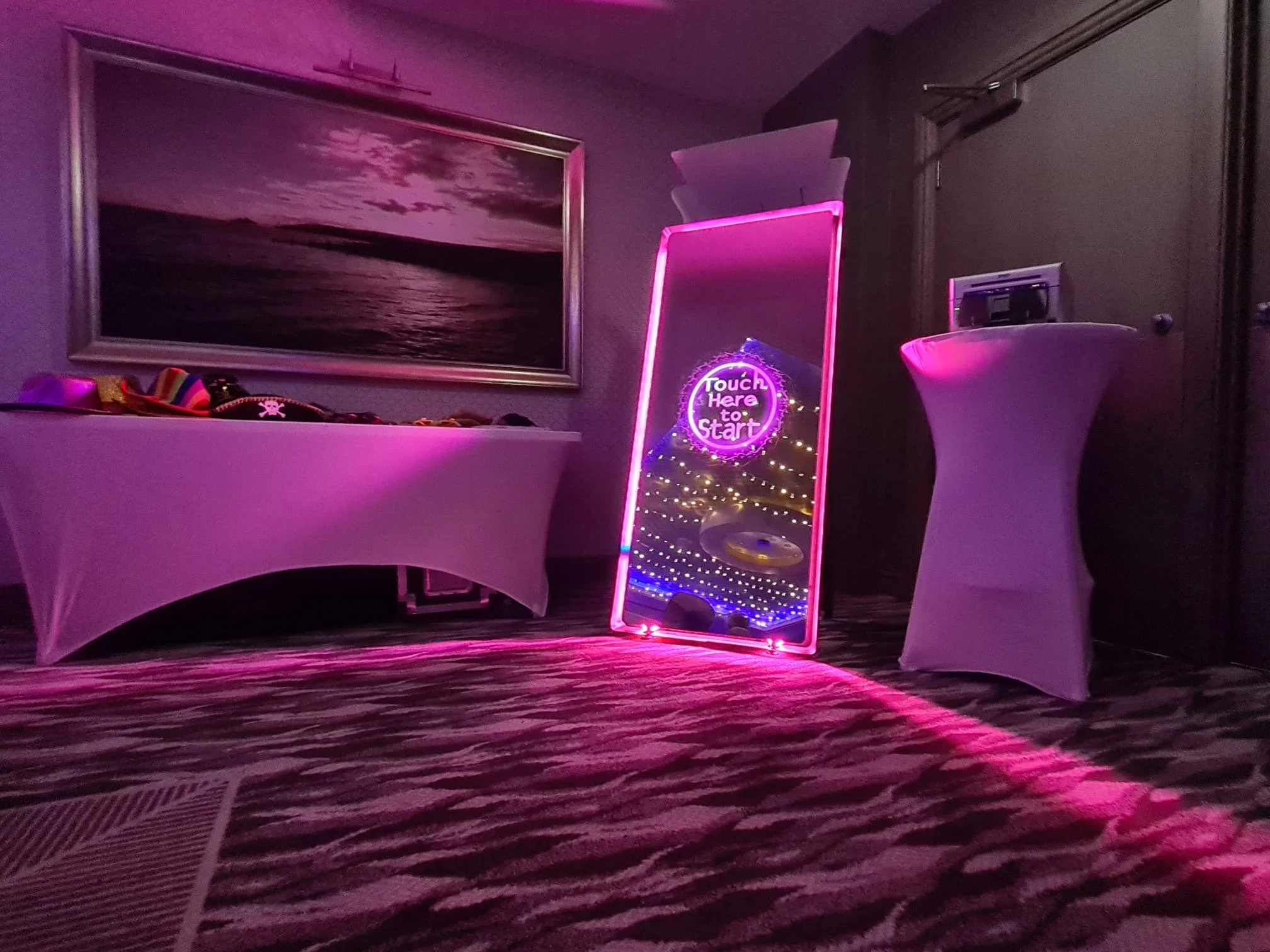 A dimly lit room with a pink and purple neon-lit 'Touch Here to Start' mirror, a table with assorted hats, a large framed landscape photograph of a sunset over water, and a high-top cocktail table. The floor has a patterned carpet, and a vent is near the floor.