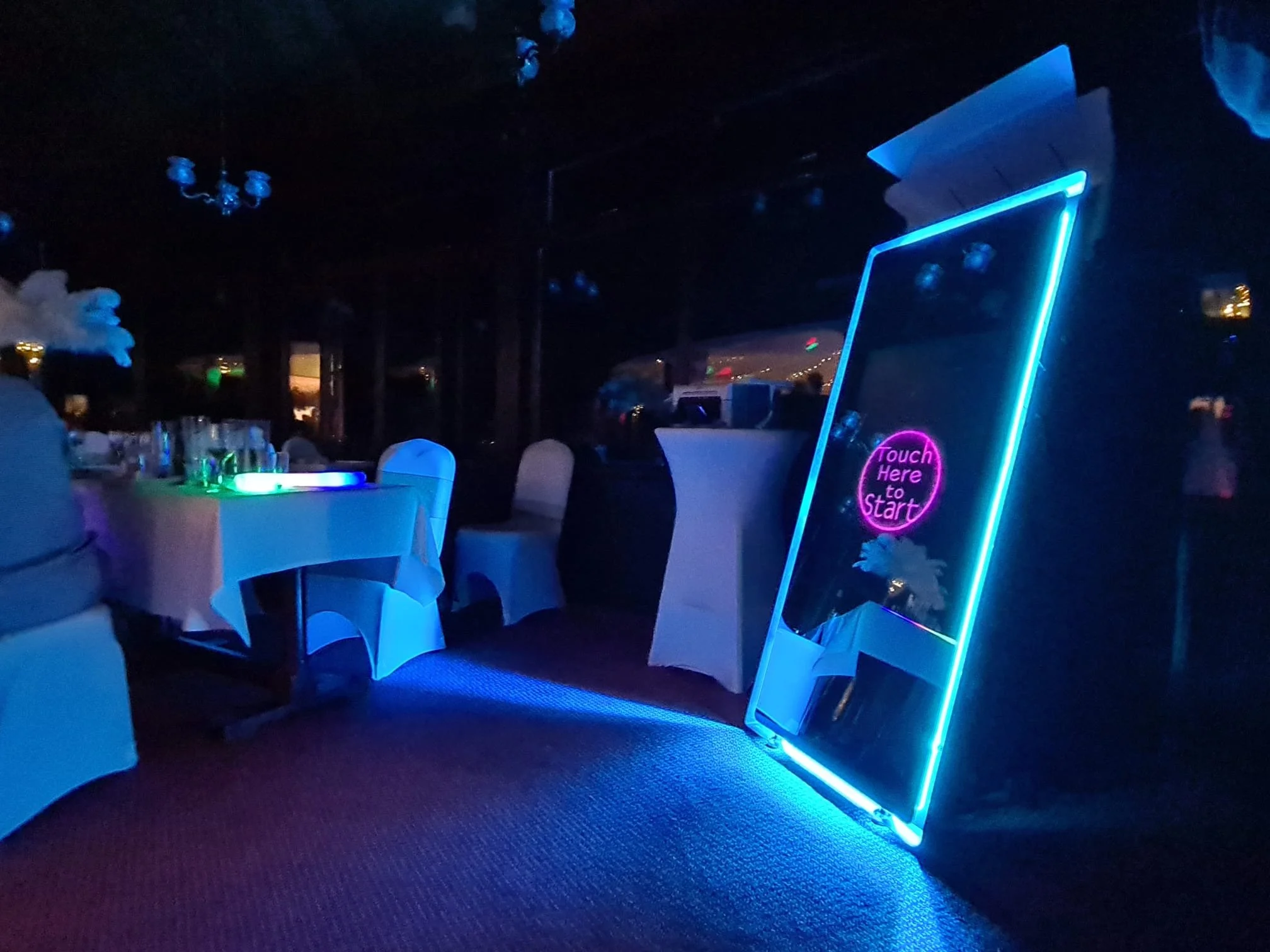 Neon-lit mirror with a glowing pink sign that says 'Touch Here to Start' in a dimly lit event space with tables and chairs.