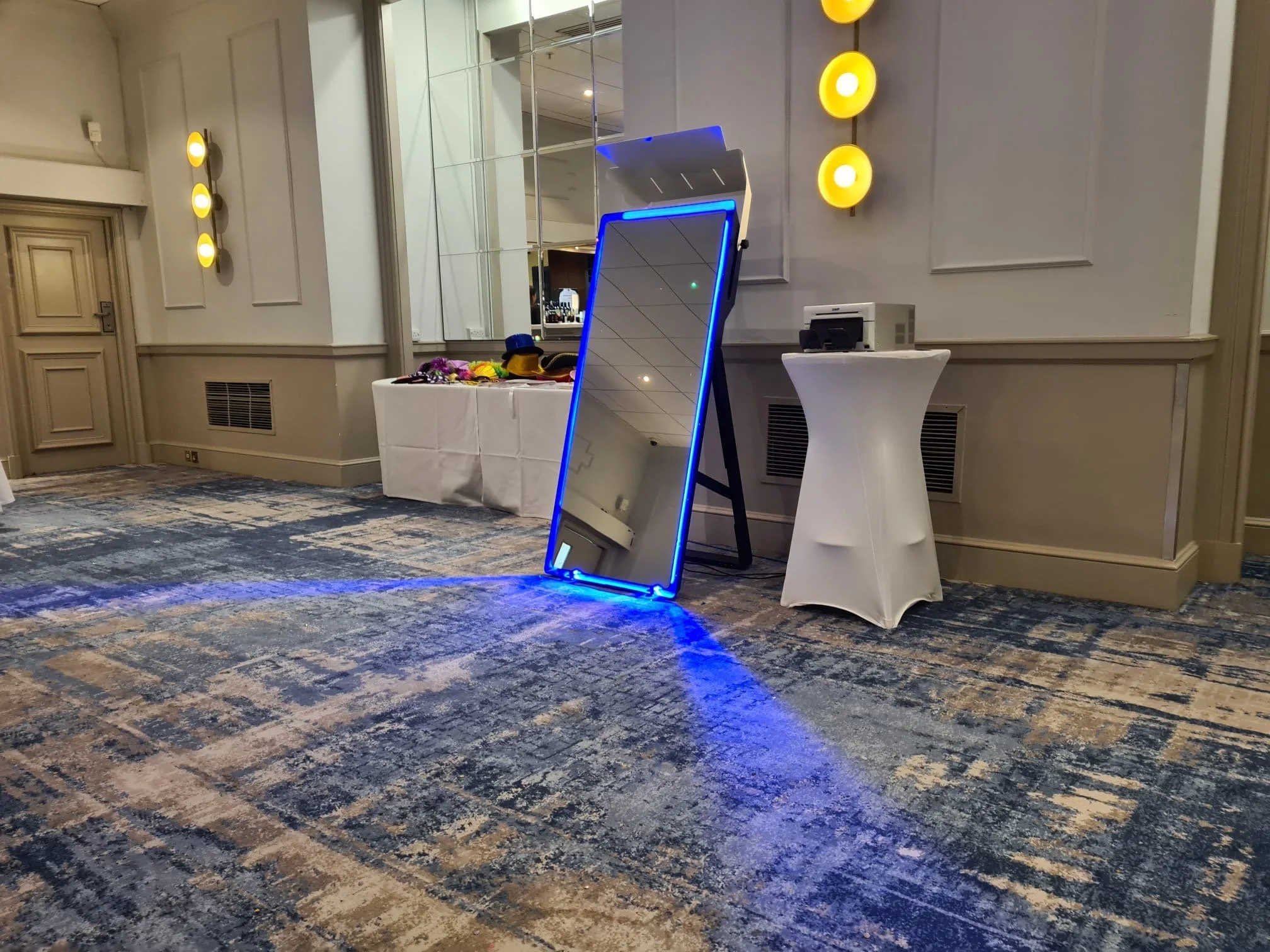 A tall rectangular mirror with blue LED lights along its edges leaning against a wall on a patterned carpeted floor, reflecting part of the ceiling. To the right, there is a cylindrical table covered with a white cloth, holding a small device. To the left, a table covered with a white cloth holds various items, including hats and accessories. The wall behind has decorative yellow circular wall sconces, and there is a large mirror reflecting part of the room.