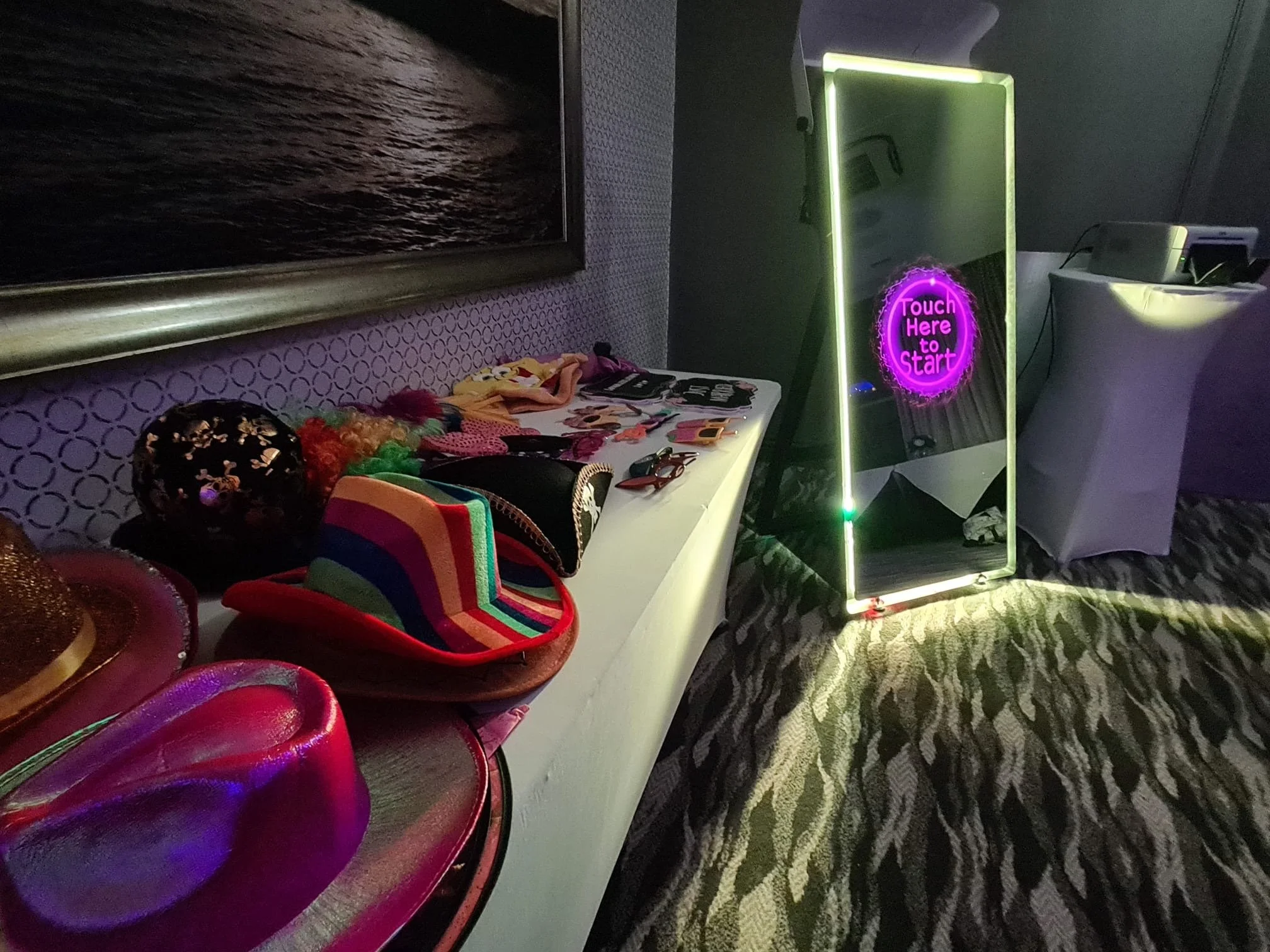 A display table with colorful party hats, wigs, and accessories, next to an illuminated mirror with a neon sign that says "Touch Here to Start." The room has patterned carpet, a framed picture on the wall, and a large purple chair.
