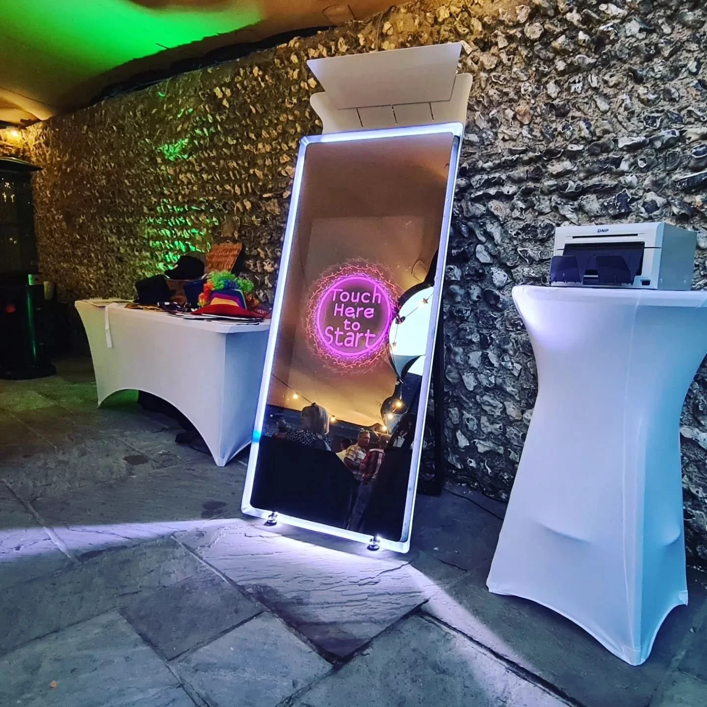 A large digital mirror with neon pink text that reads "Touch Here to Start" leaning against a stone wall at an event. There are two white high-top tables nearby, one with a printer and the other with various items including hats and colorful accessories. Reflections of people and decorative lighting are visible in the mirror.