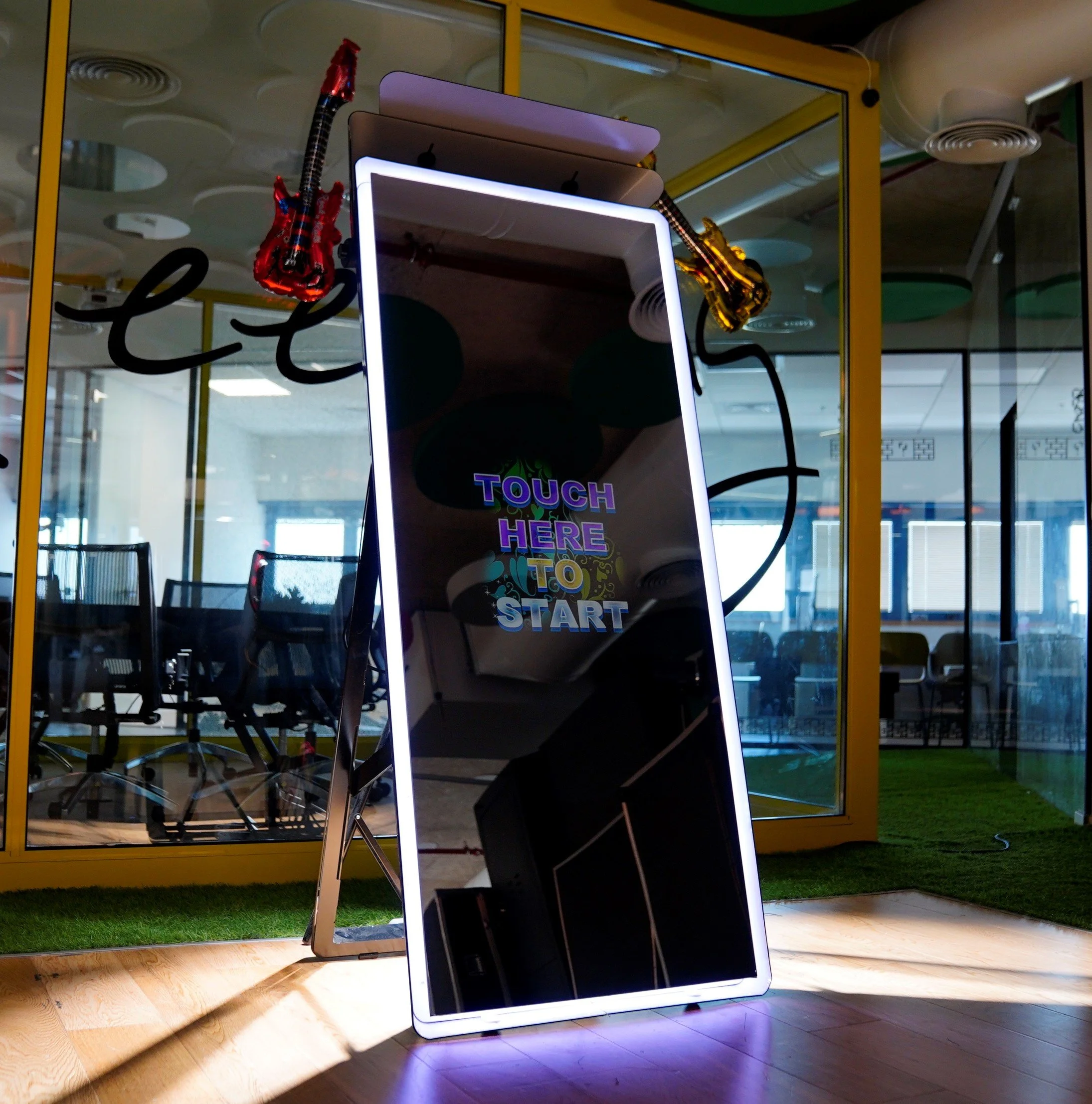A large, upright mirror with the words 'TOUCH HERE TO START' illuminated on it, situated in a modern office or event space with glass walls and chairs in the background.