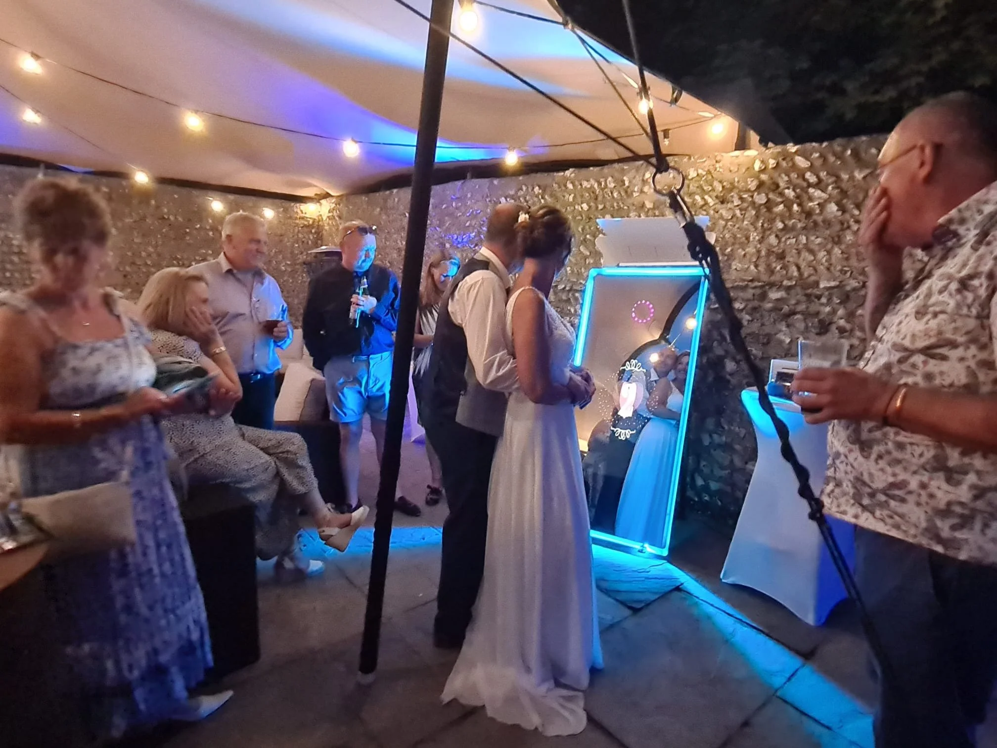 People attending a party or celebration in an outdoor space with string lights, a stone wall, a photo booth with a Disney princess design, and some individuals standing and chatting.