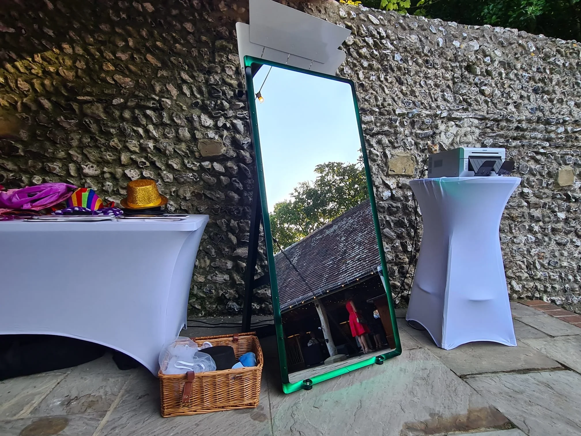 Outdoor event setup with a large free-standing mirror reflecting a person in a red dress, two tall tables with white tablecloths holding various items, a wicker basket with miscellaneous items, and a stone wall background.