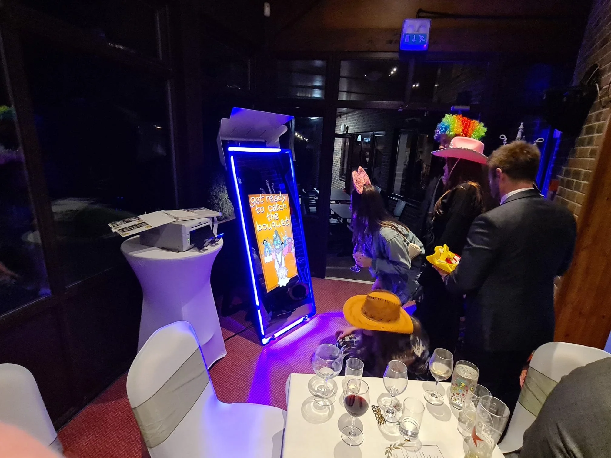 People dressed in colorful costumes and hats standing in line at a photo booth with a bright digital sign reading "get ready to catch the bouquet" in a room with dark walls and tables with glasses and drinks.