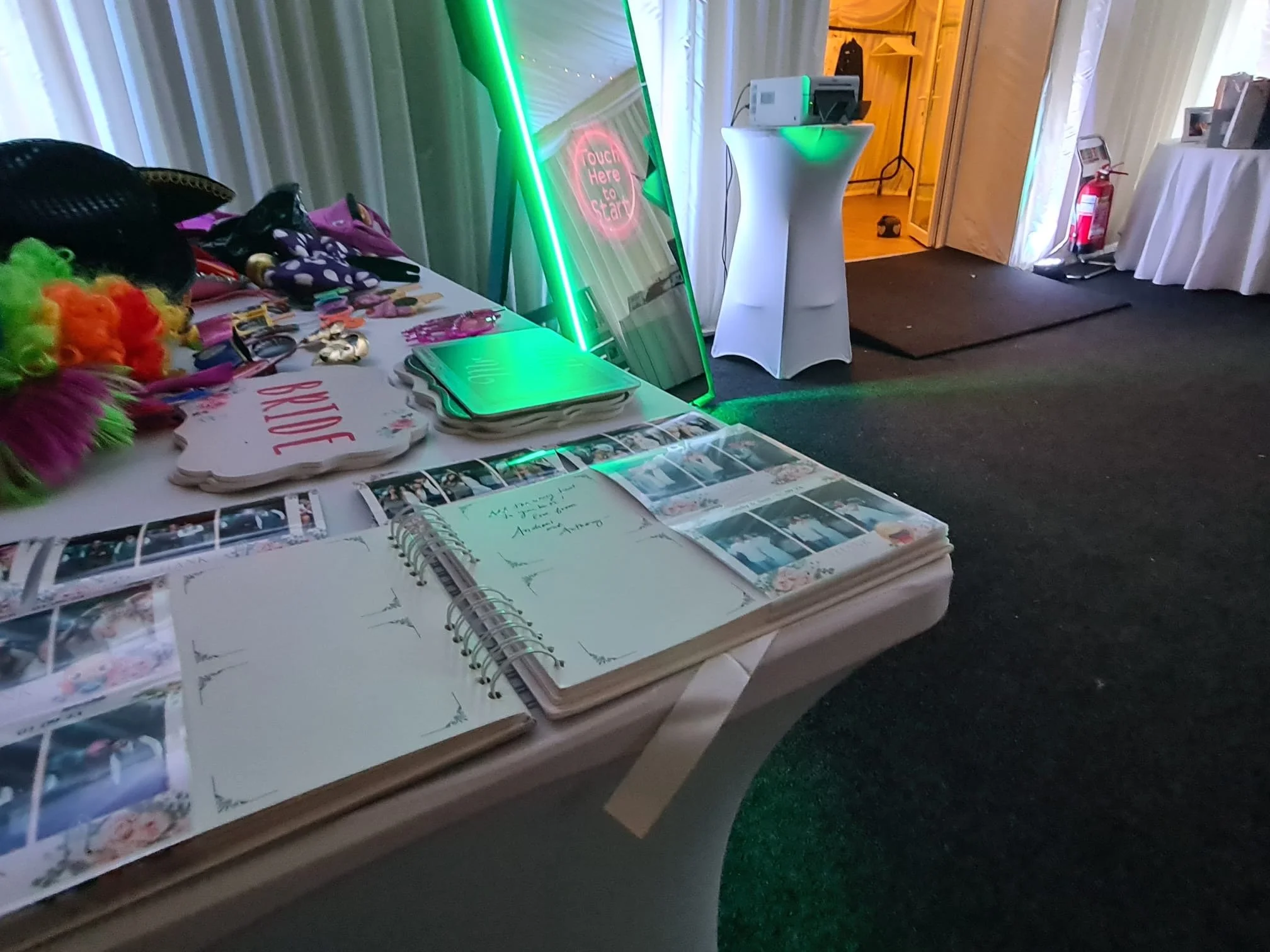 Photo of a display table with colorful props, photo booth pictures, and a sign that says "LOVE". A neon-lit mirror with "Touch Here to Start" is leaning against the wall next to a small round table with a printer and a green-lit lamp. An open guest book with handwritten messages is on the table. In the background, there is a doorway, a flickering light, and a fire extinguisher.