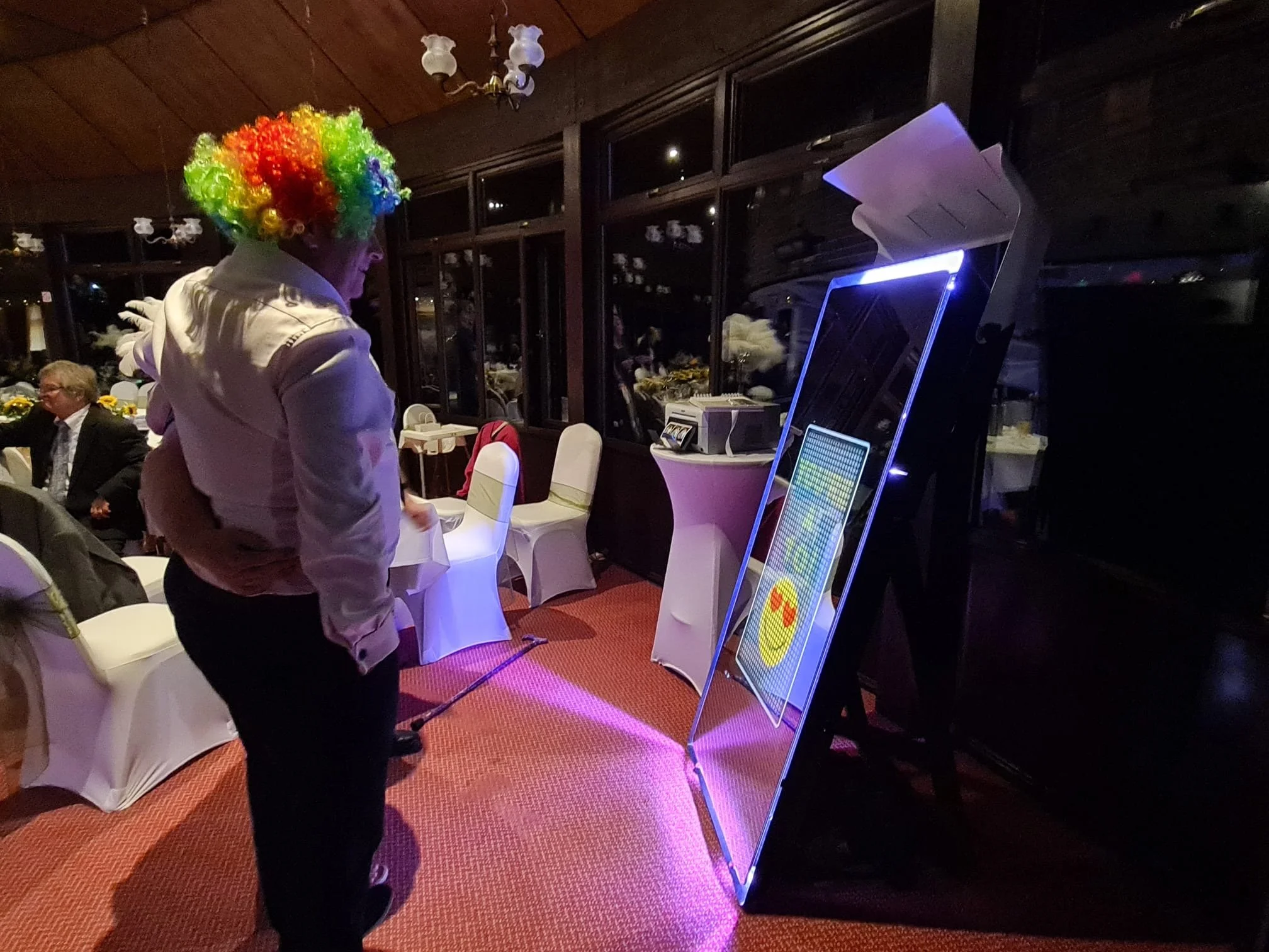 A man wearing a colorful clown wig and white jacket stands in a dimly lit room, looking at an illuminated photo booth or digital display.