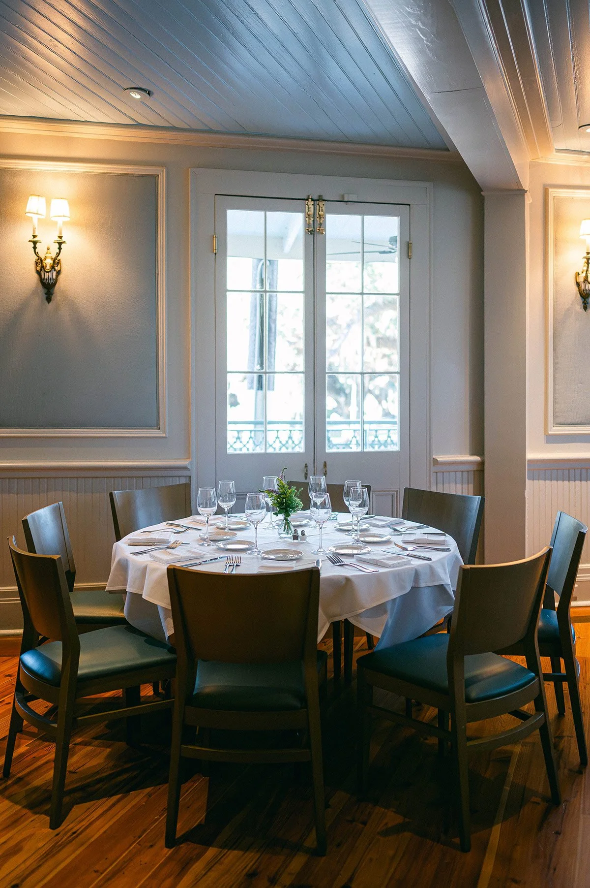 A round dining table set with white tablecloth, glassware, plates, and silverware, in a well-lit room with wood flooring, wall sconces, and French doors leading outside.
