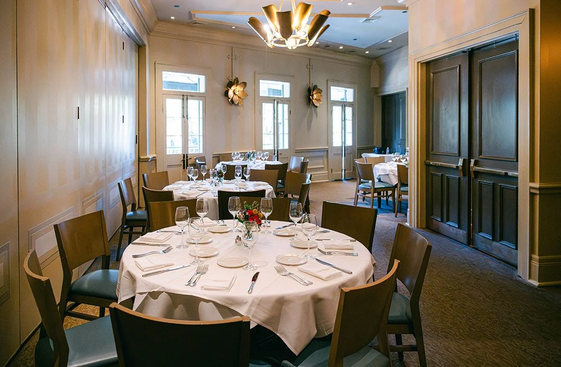 Elegant dining room with round tables set for a formal meal, featuring white tablecloths, glassware, and silverware, with windows, wall decorations, and a chandelier.