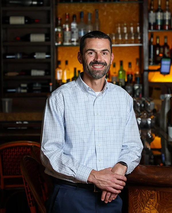 Man with a beard in a white checkered shirt standing behind a bar with liquor bottles in the background.