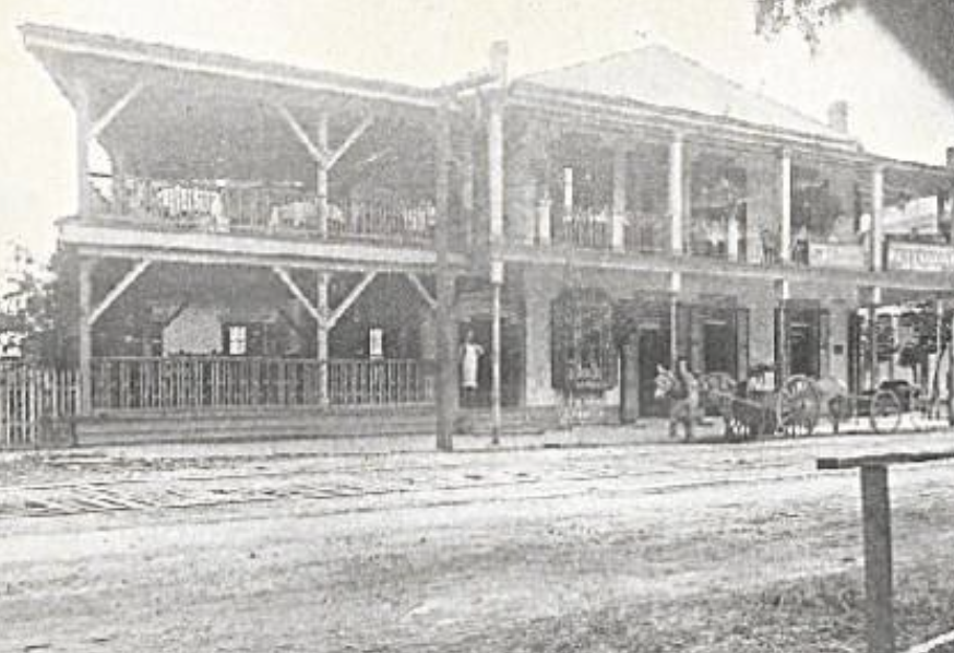 Black and white photo of Ralph's on the Park in 1860 when it was a coffeeshop, a two-story wooden building with a porch on each level, a person standing on the sidewalk, and a horse-drawn carriage parked in front.