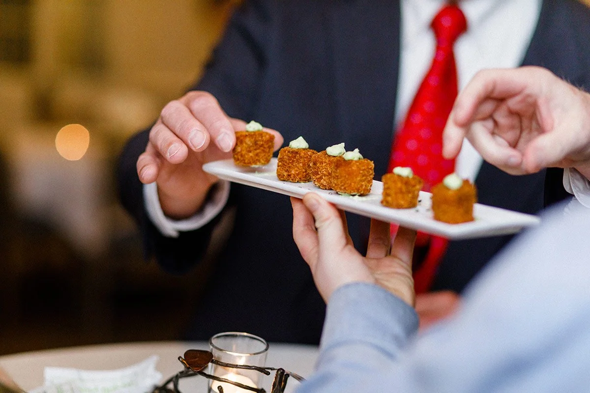 Person in a suit and red tie receiving a plate of small, breaded, fried appetizers topped with a dollop of white sauce.