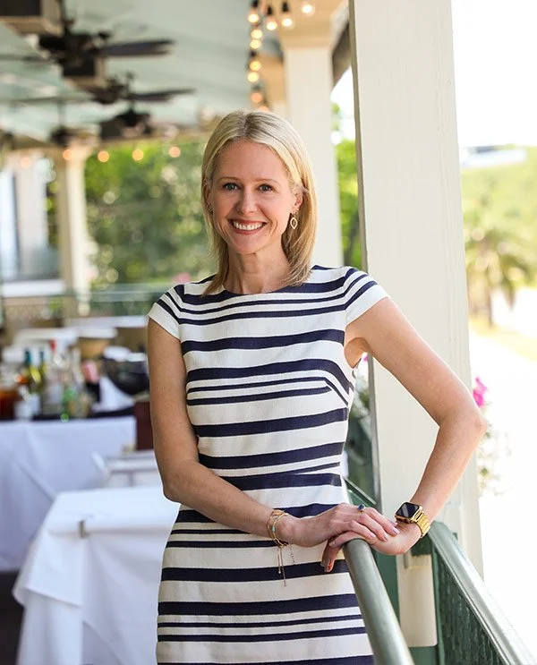 A smiling woman in a white and navy striped dress standing outdoors on a balcony or patio area, with a blurred background of trees and outdoor furniture.