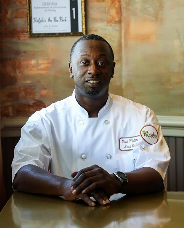 A man in a white chef's coat sitting at a dining table in a restaurant, with a name tag that reads 'Sam Winters,' and a Ralph's badge on his coat.