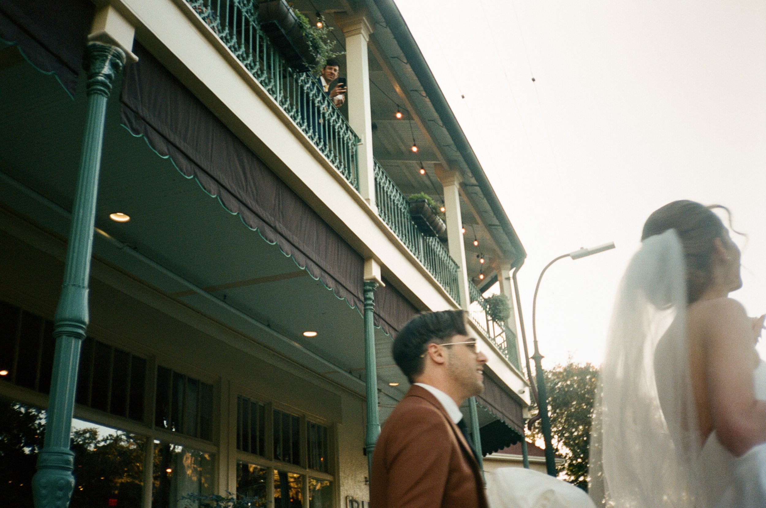 A wedding scene showing a bride with veil and a groom in a brown suit walking outdoors near a two-story building with a balcony and decorative railings, with a person taking photos on the balcony.