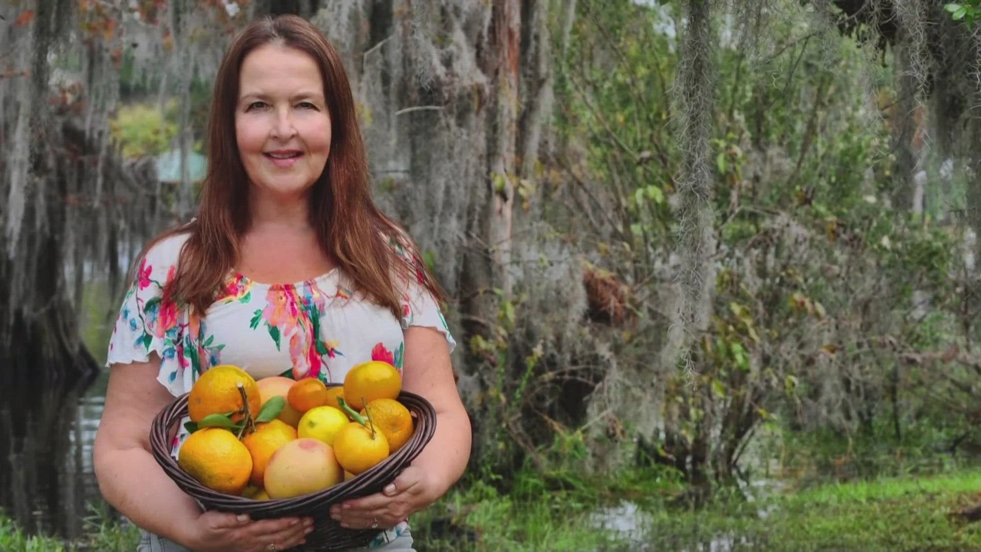 A woman with brown hair, wearing a floral top, holding a basket filled with various apples and oranges, standing outdoors in front of a swampy area with trees and Spanish moss hanging from branches.