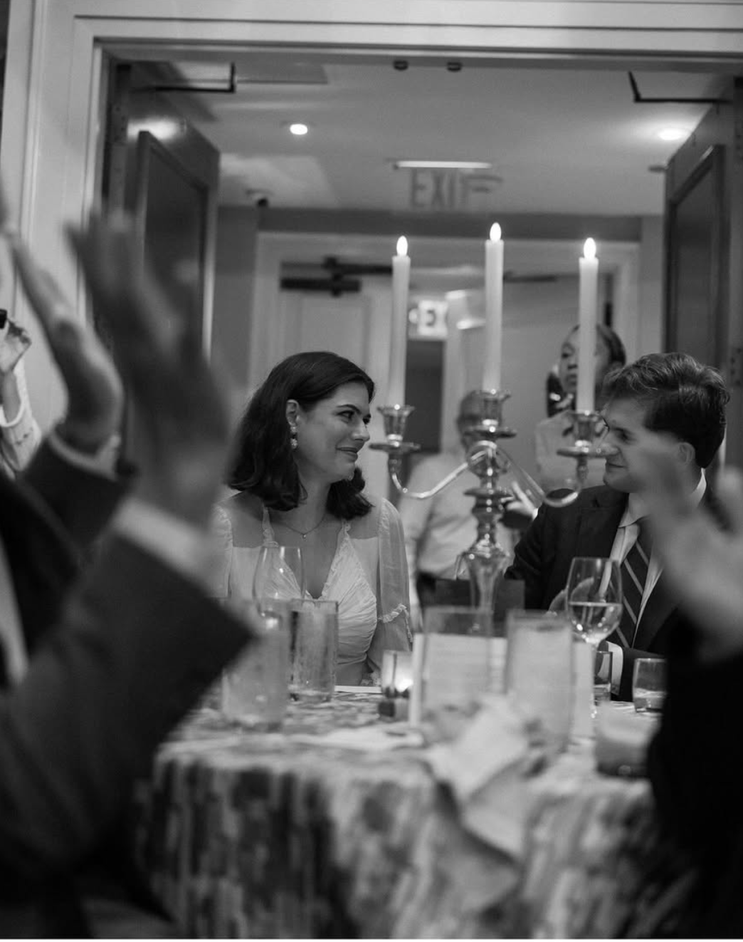 People sitting at a banquet table with lit candles, a woman with dark hair smiling at a man in a suit, in a formal setting.