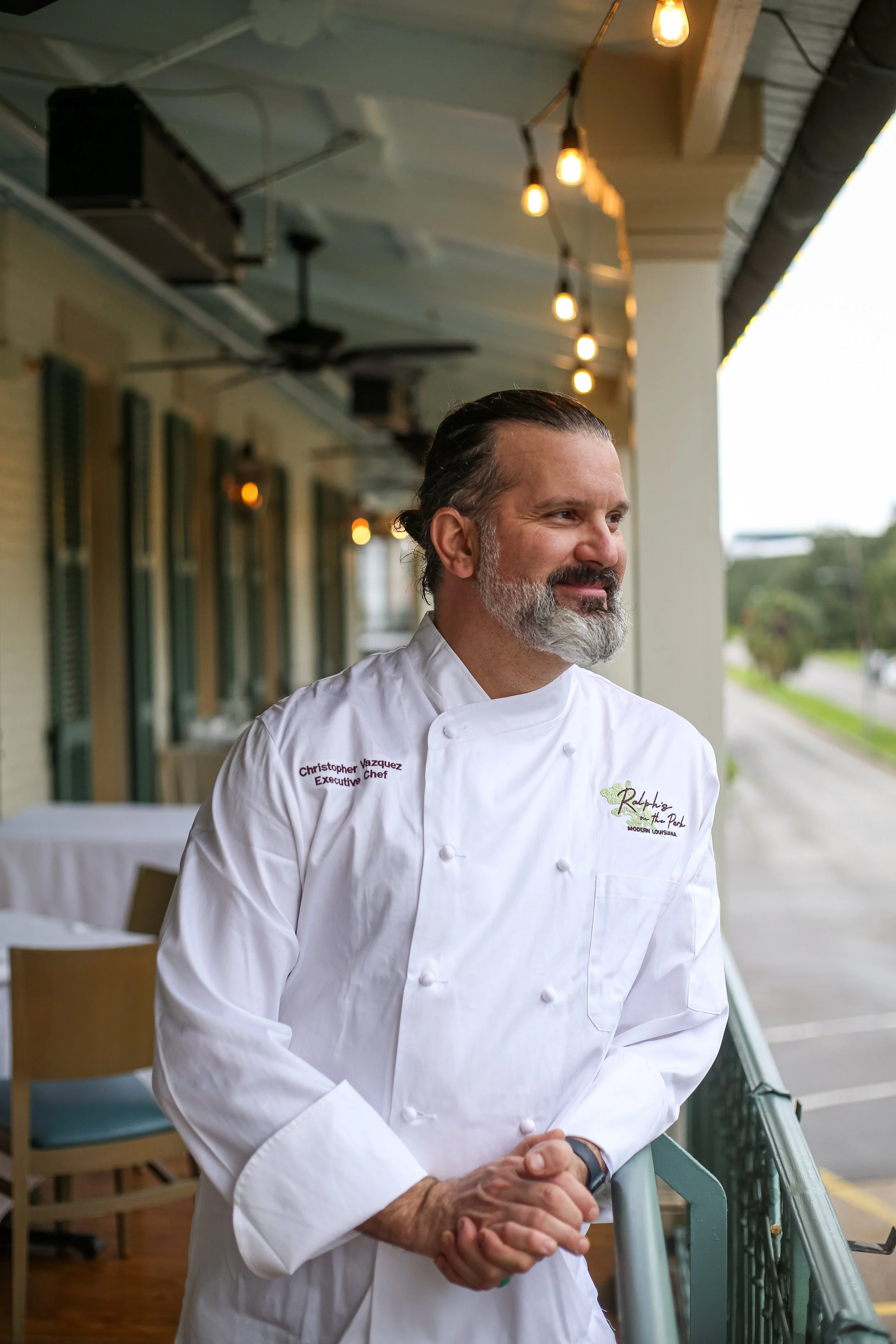 A man dressed in a white chef's coat standing on a balcony, looking off to the side with a slight smile, with a street in the background.