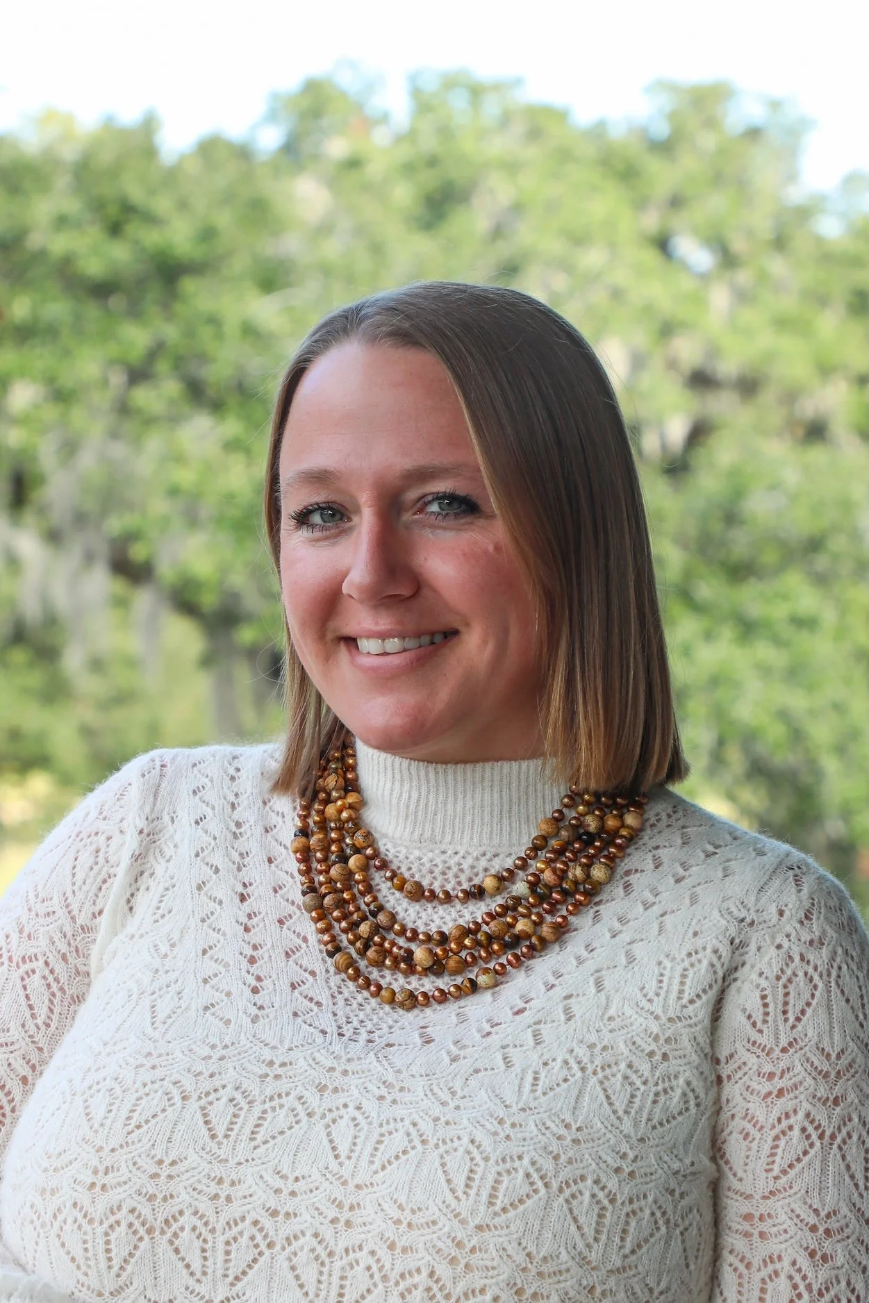 A woman with shoulder-length brown hair, wearing a cream lace sweater and layered beaded necklaces, smiling outdoors with green trees in the background.