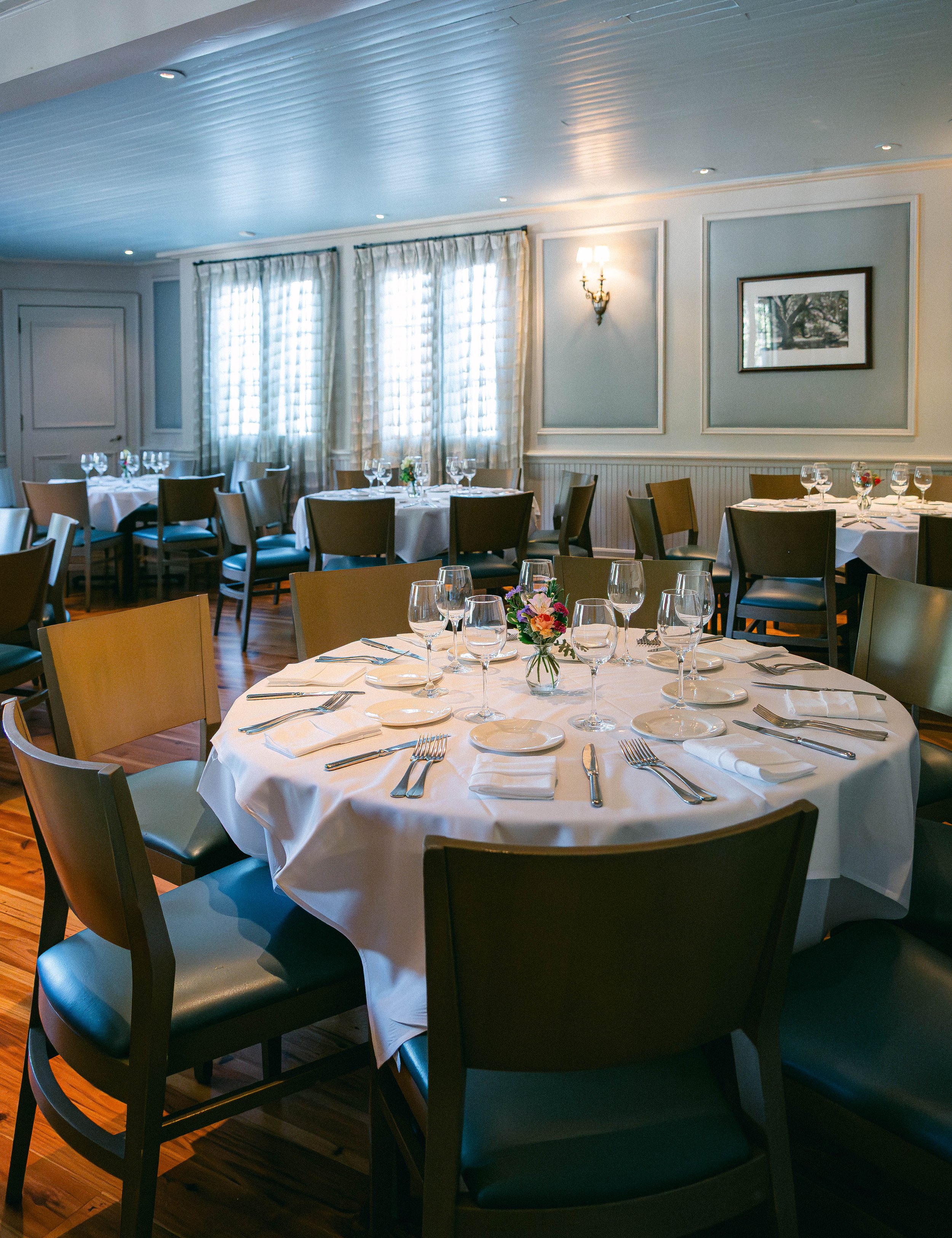 A round dining table set with white tablecloth, glassware, plates, and silverware, in a well-lit room with wood flooring, wall sconces, and French doors leading outside.