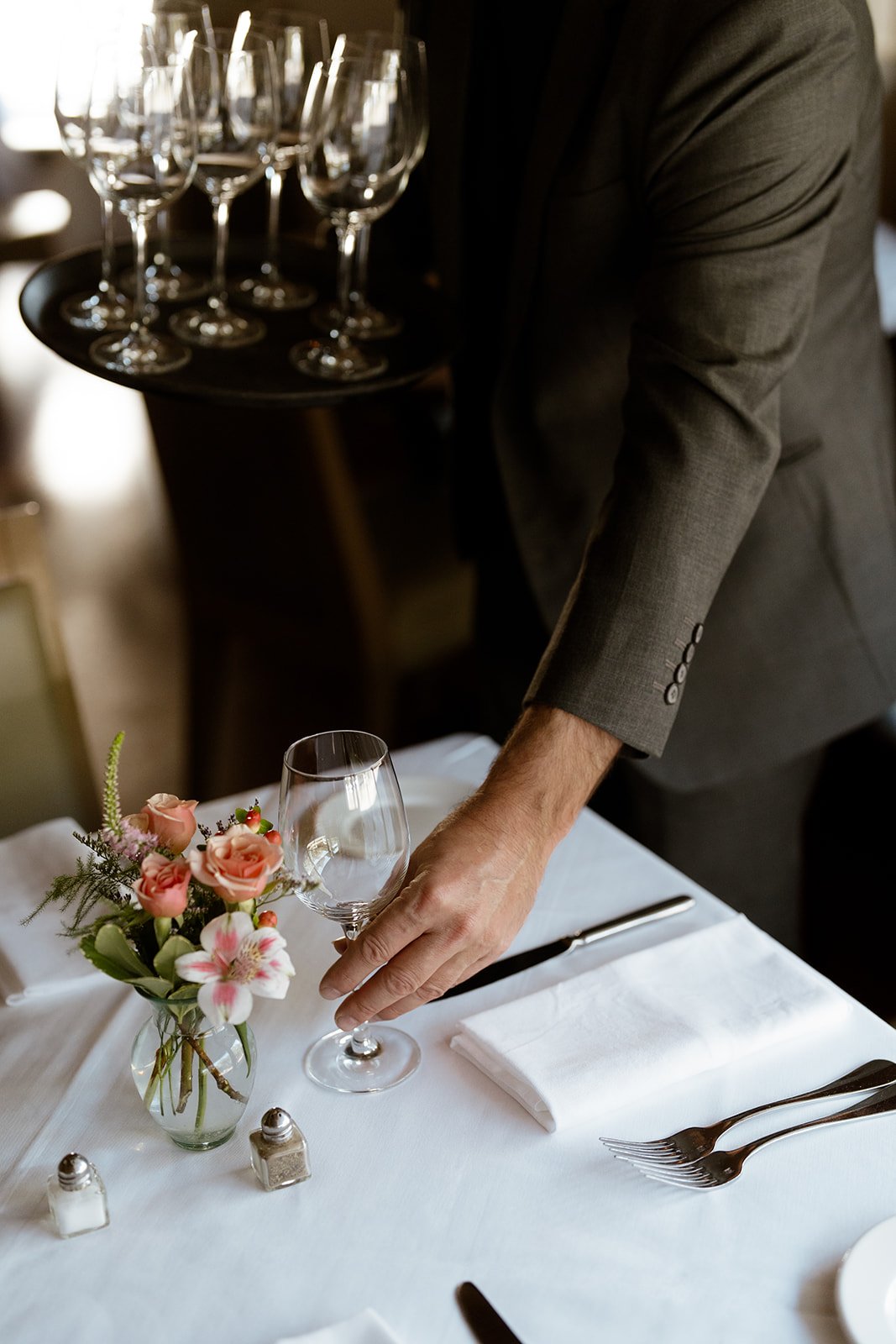 A waiter in a gray suit setting a wine glass on a white tablecloth with a flower arrangement.