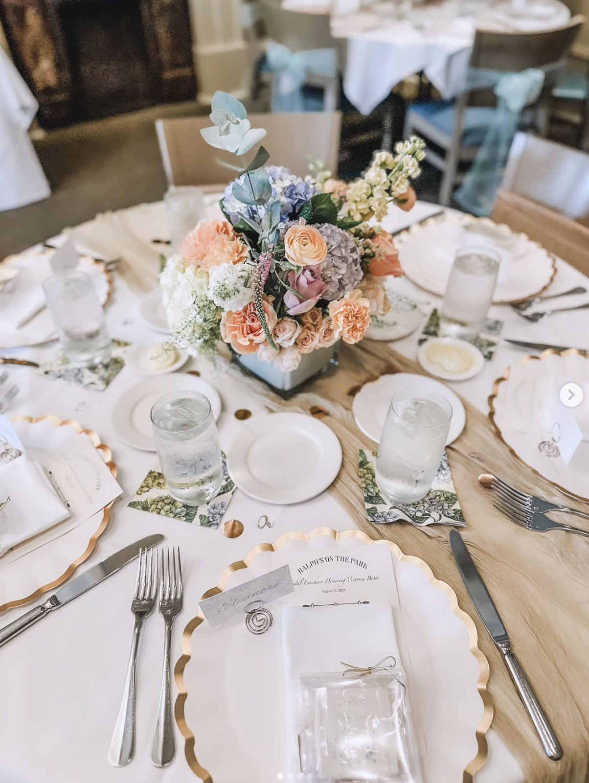 A round wedding reception table decorated with a floral centerpiece, white plates with gold scalloped edges, silverware, water glasses, and printed menus.