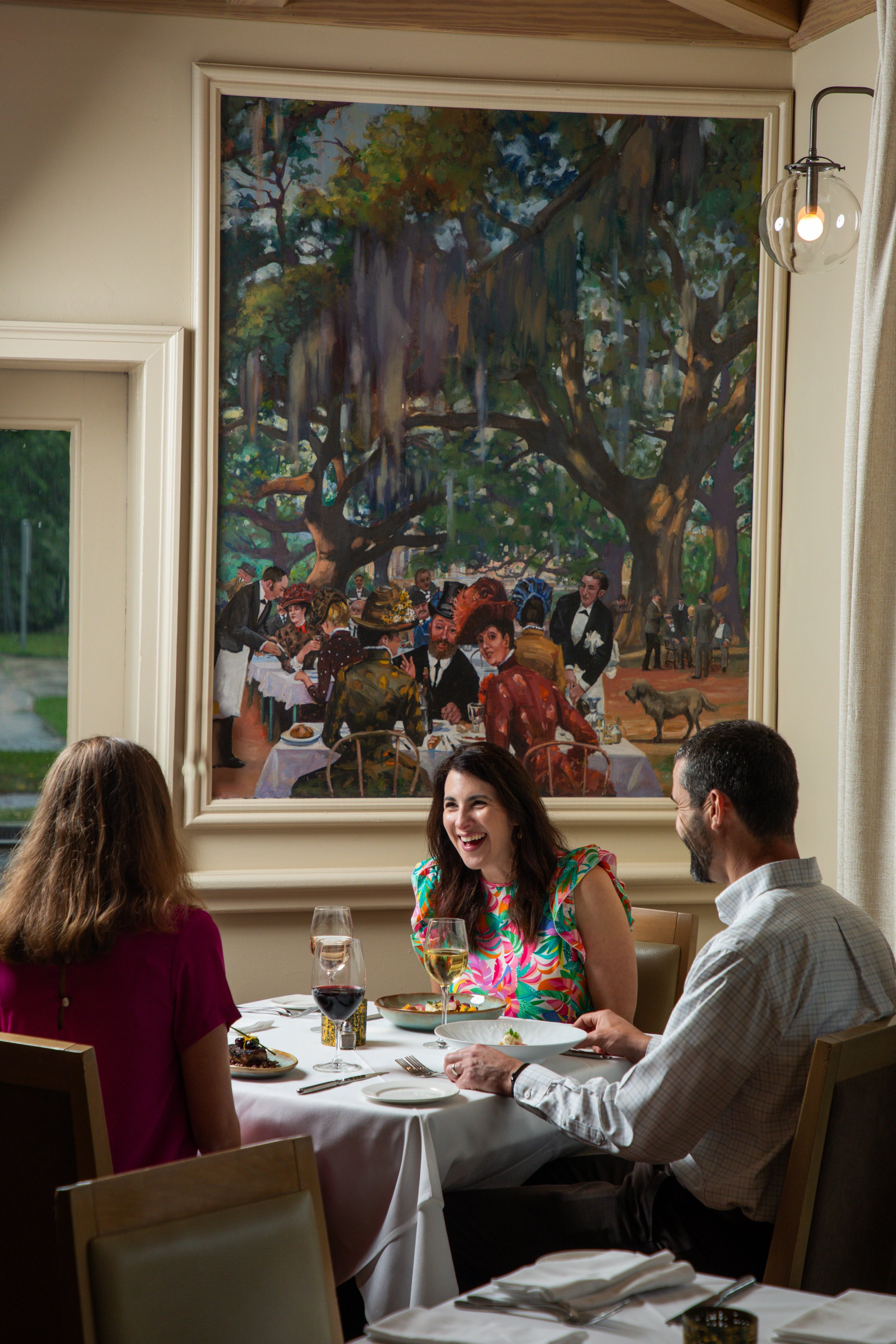 People dining at a restaurant table with a large indoor painting of an outdoor scene with trees and people in the background.