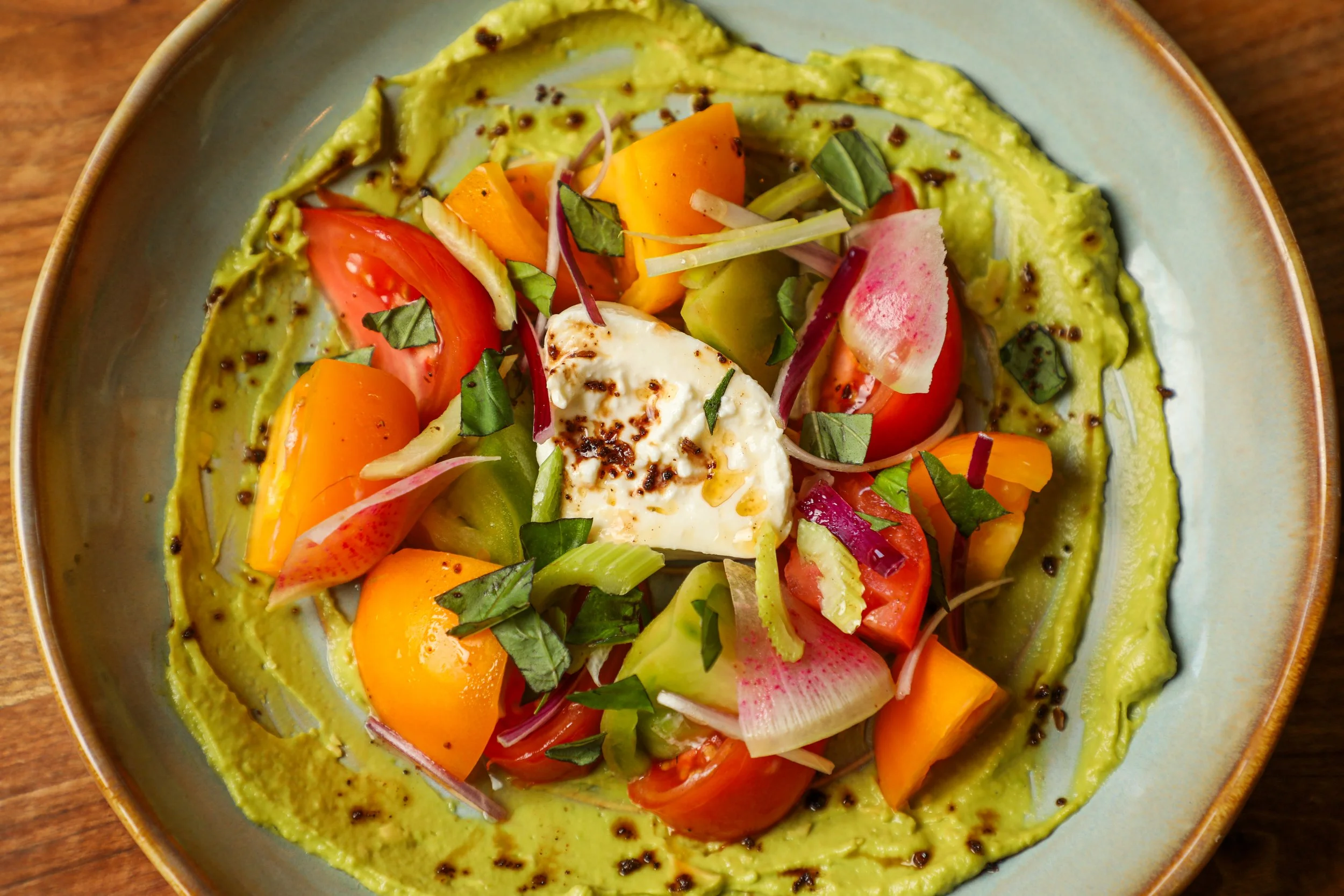 A plate of colorful salad with diced tomatoes, sliced radishes, yellow and orange bell peppers, green cucumber, avocado, topped with a poached egg and chopped basil, surrounded by a drizzled green sauce.