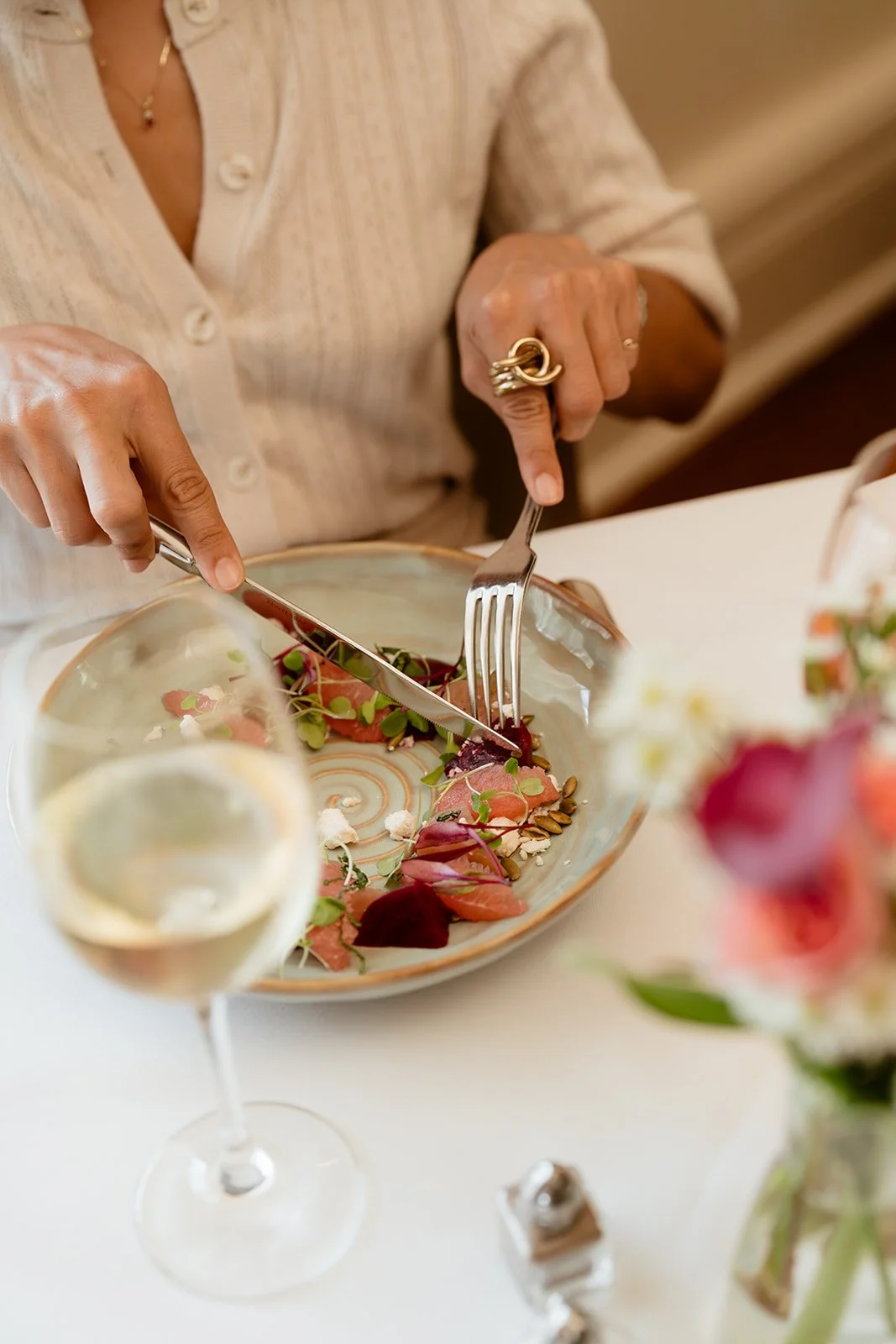 Person cutting a dish of smoked salmon garnished with microgreens and edible flowers, with a glass of white wine nearby.