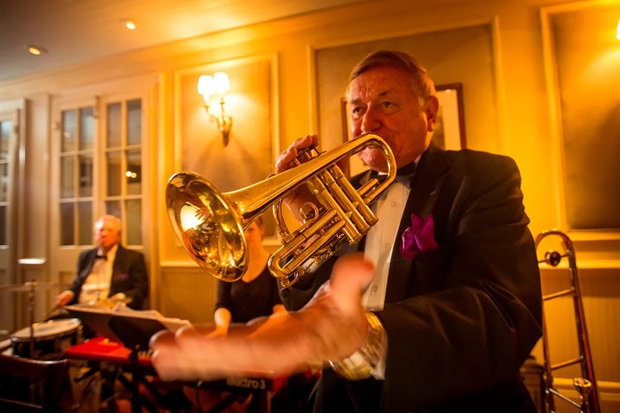 Man in tuxedo playing a trumpet at a formal event with warm lighting and elegant decor.