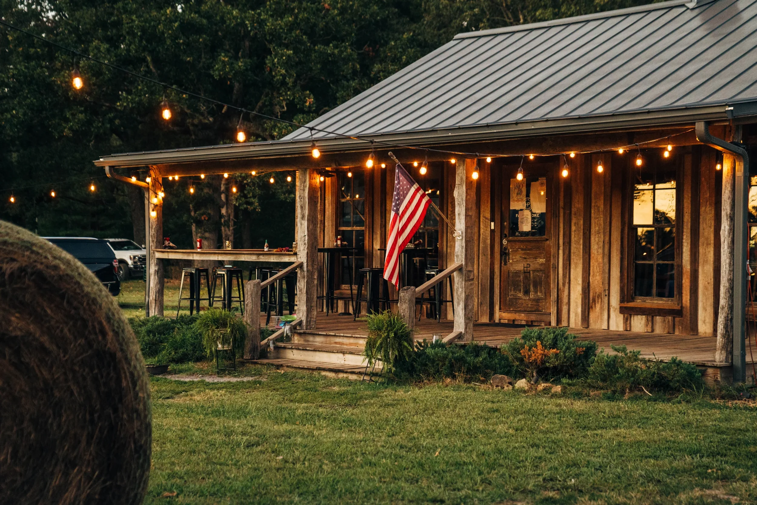 A rustic wooden house decorated with string lights and an American flag on the front porch, surrounded by greenery, with parked cars in the background during evening