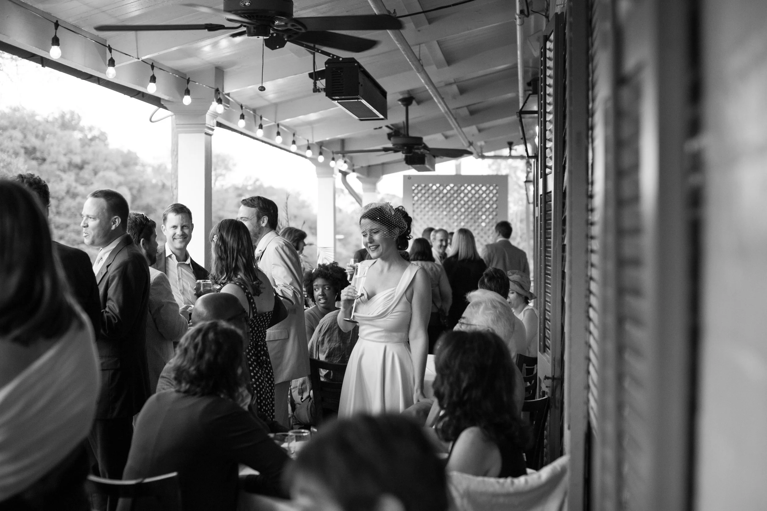 People at an outdoor wedding reception, some are sitting and some are standing, with a woman in a white dress holding a drink and smiling.