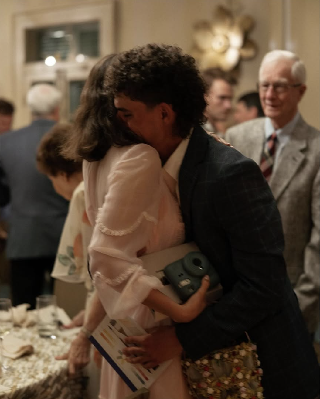 Two women hugging at a social gathering, with one holding a camera and a book, and older man smiling in the background.