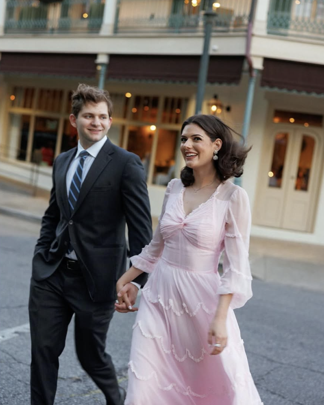 A smiling couple walking hand in hand outside a building, dressed in formal attire, with the woman in a light pink dress and the man in a dark suit with a striped tie.