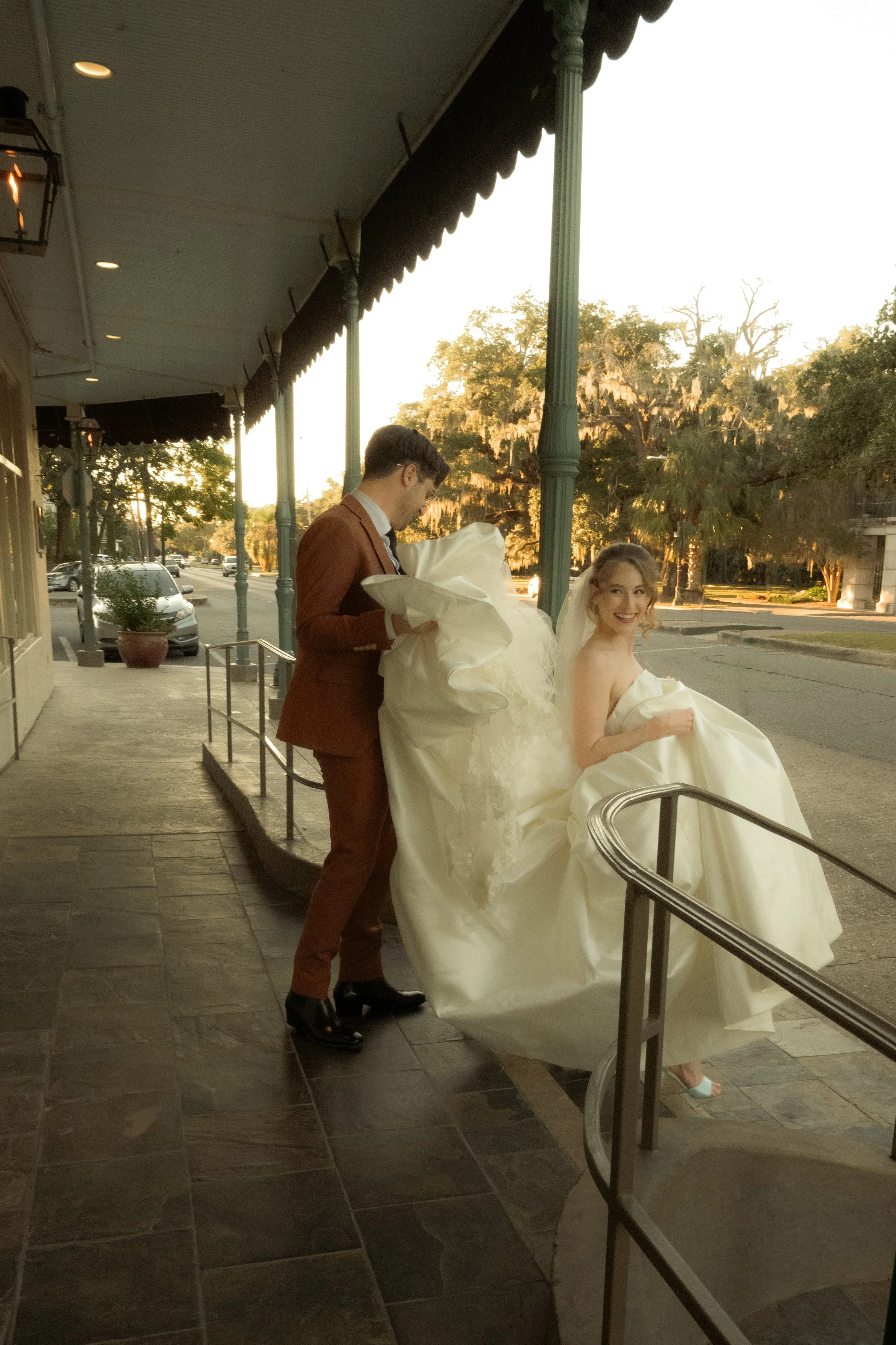 A newlywed couple, the bride in a wedding gown sitting on a railing and the groom in a suit standing beside her, outside on a sidewalk during sunset.