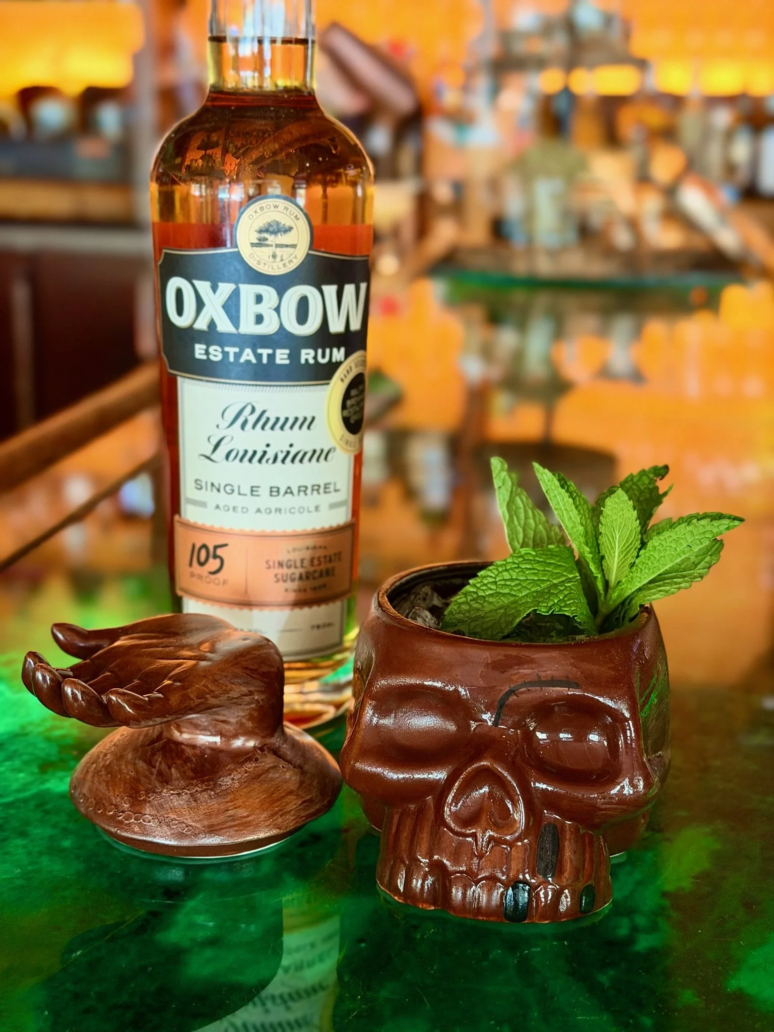Ceramic tiki mug with a face design, featuring pink floral decorations around the top, and a straw in the mug, set against a blurred outdoor background.